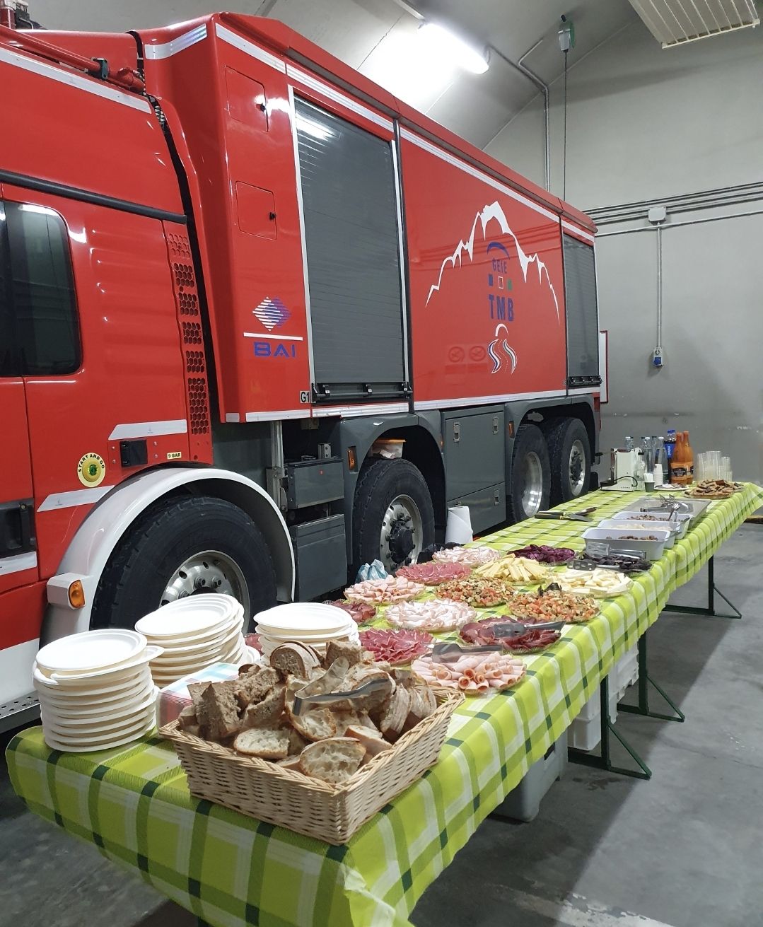 Un lungo tavolo da buffet con pane, salumi e pile di piatti, parcheggiato accanto a un grande veicolo rosso di pronto intervento.