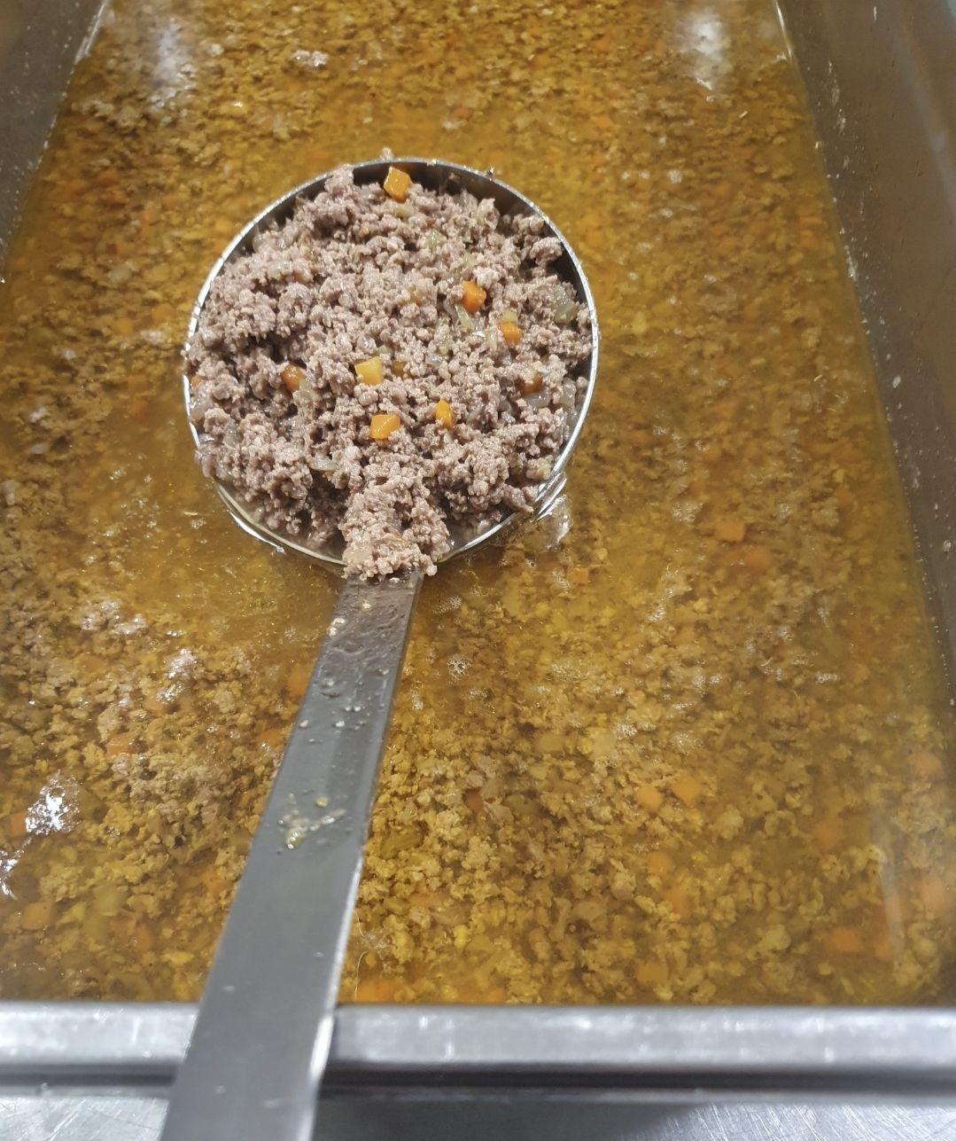 Un mestolo di metallo pieno di carne macinata e piccoli pezzi di verdure arancioni, sollevato da una grande pentola di sugo di carne ricco di liquido.