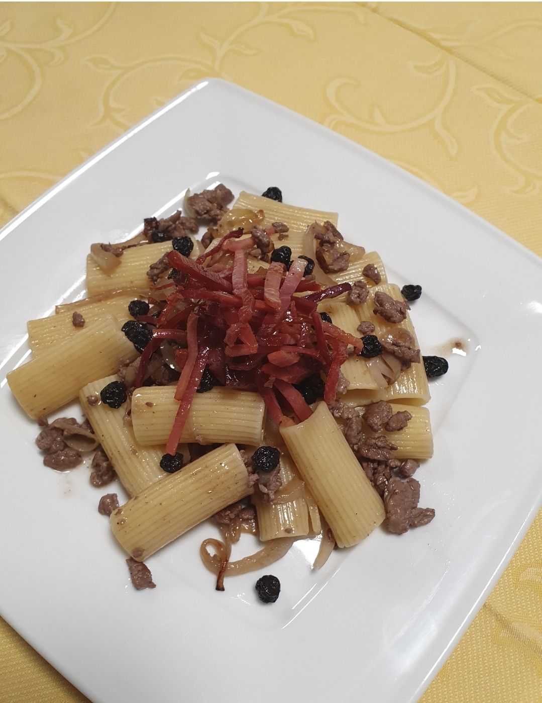 Un piatto di rigatoni conditi con carne macinata, pancetta e frutti di bosco su una tovaglia gialla a motivi geometrici.