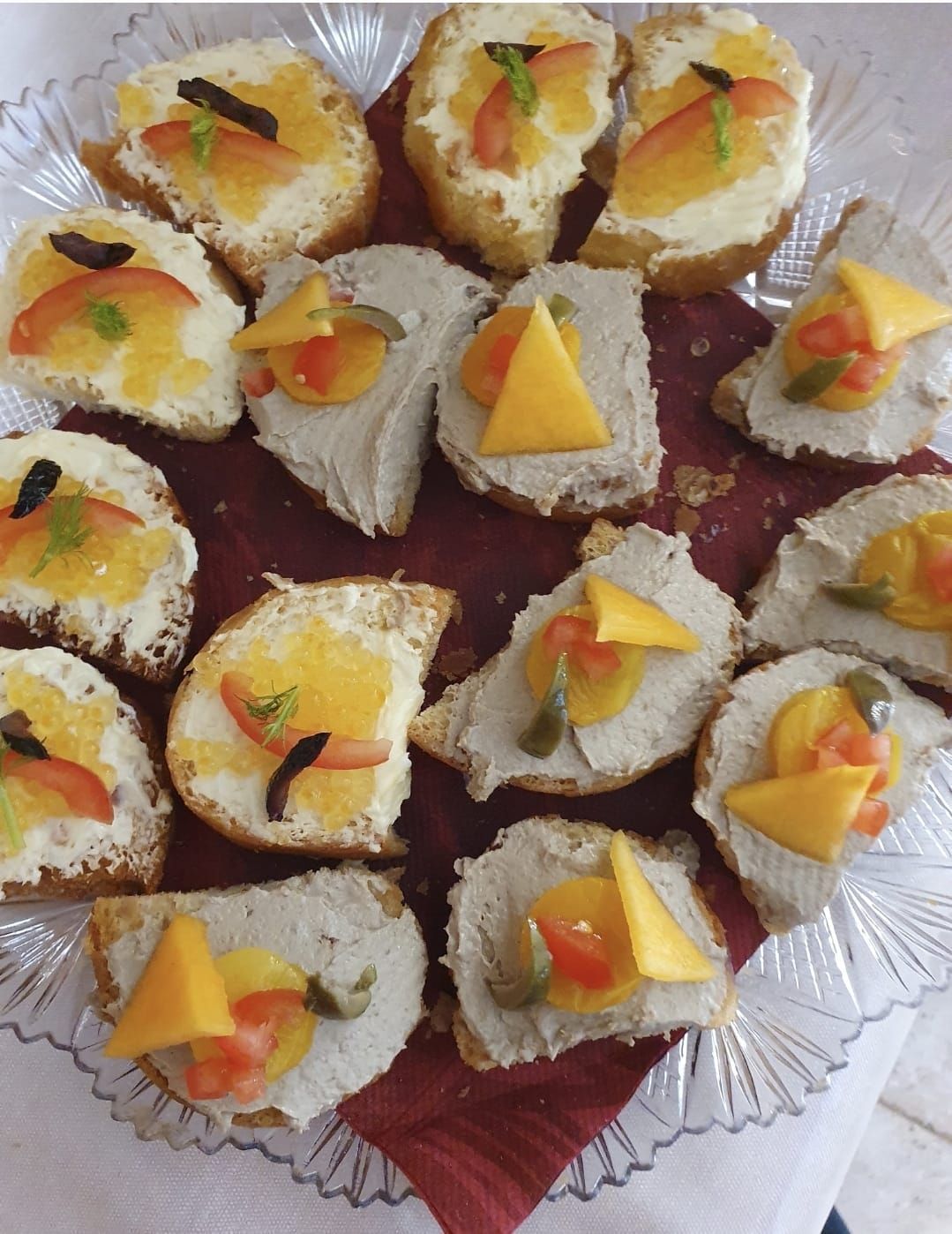 Un piatto di vetro contenente vari antipasti a base di pane tostato guarnito con formaggio bianco o paté e frutta.