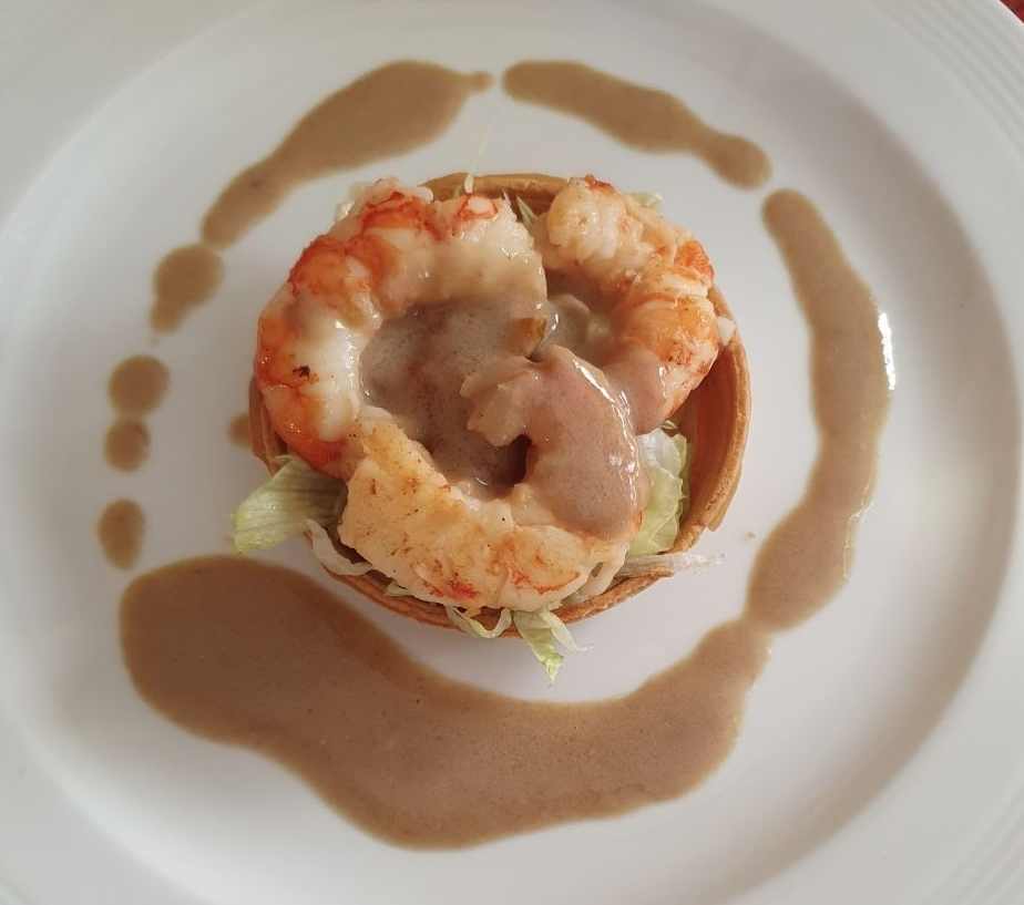 Un gambero cotto all'interno di una piccola tortina di pasta sfoglia, guarnito con lattuga e un anello di salsa bruna su un piatto bianco.