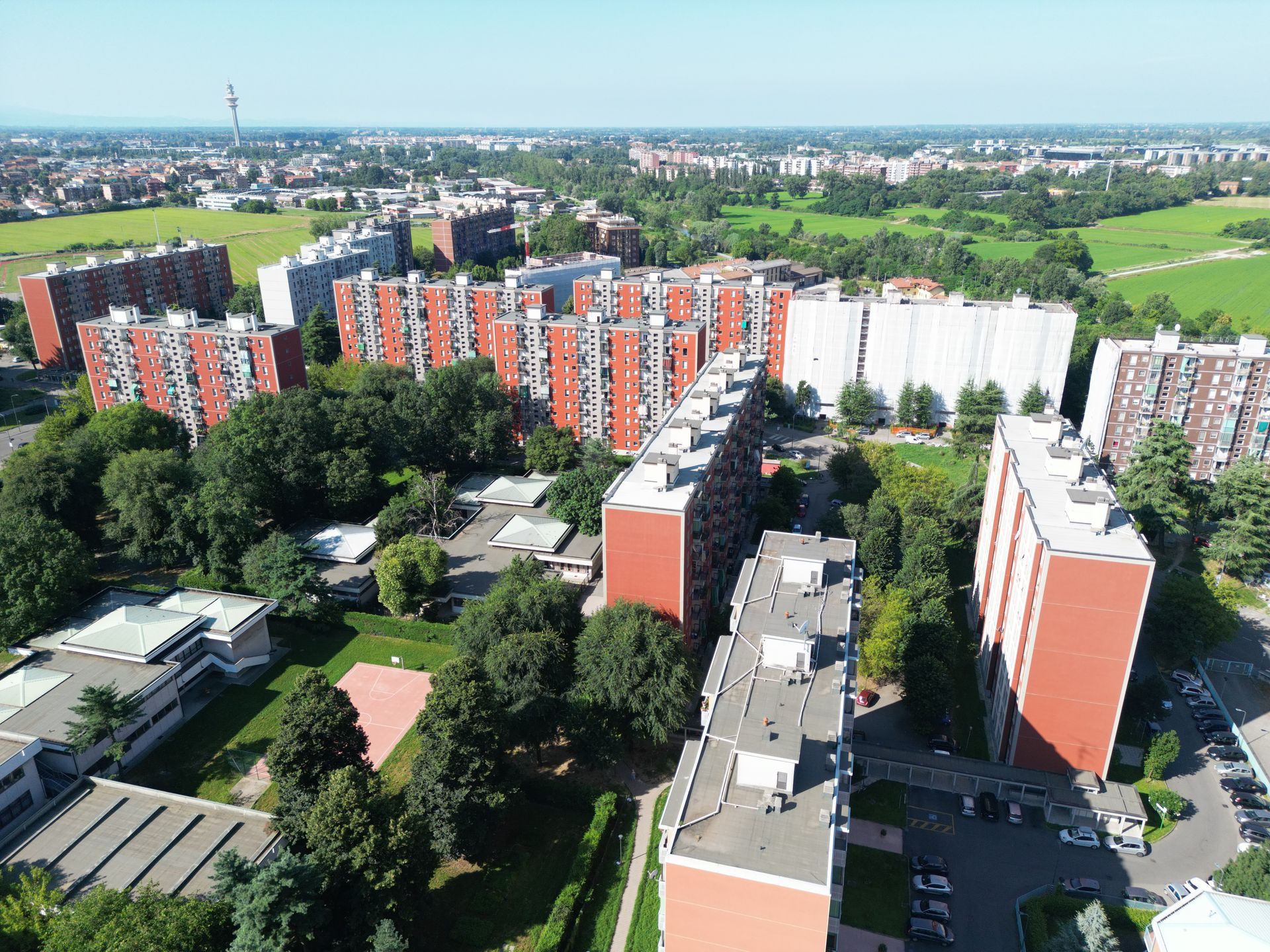 Vista aerea di diversi edifici residenziali in mattoni rossi con alberi verdi e campi, in una zona urbana in una giornata di sole.