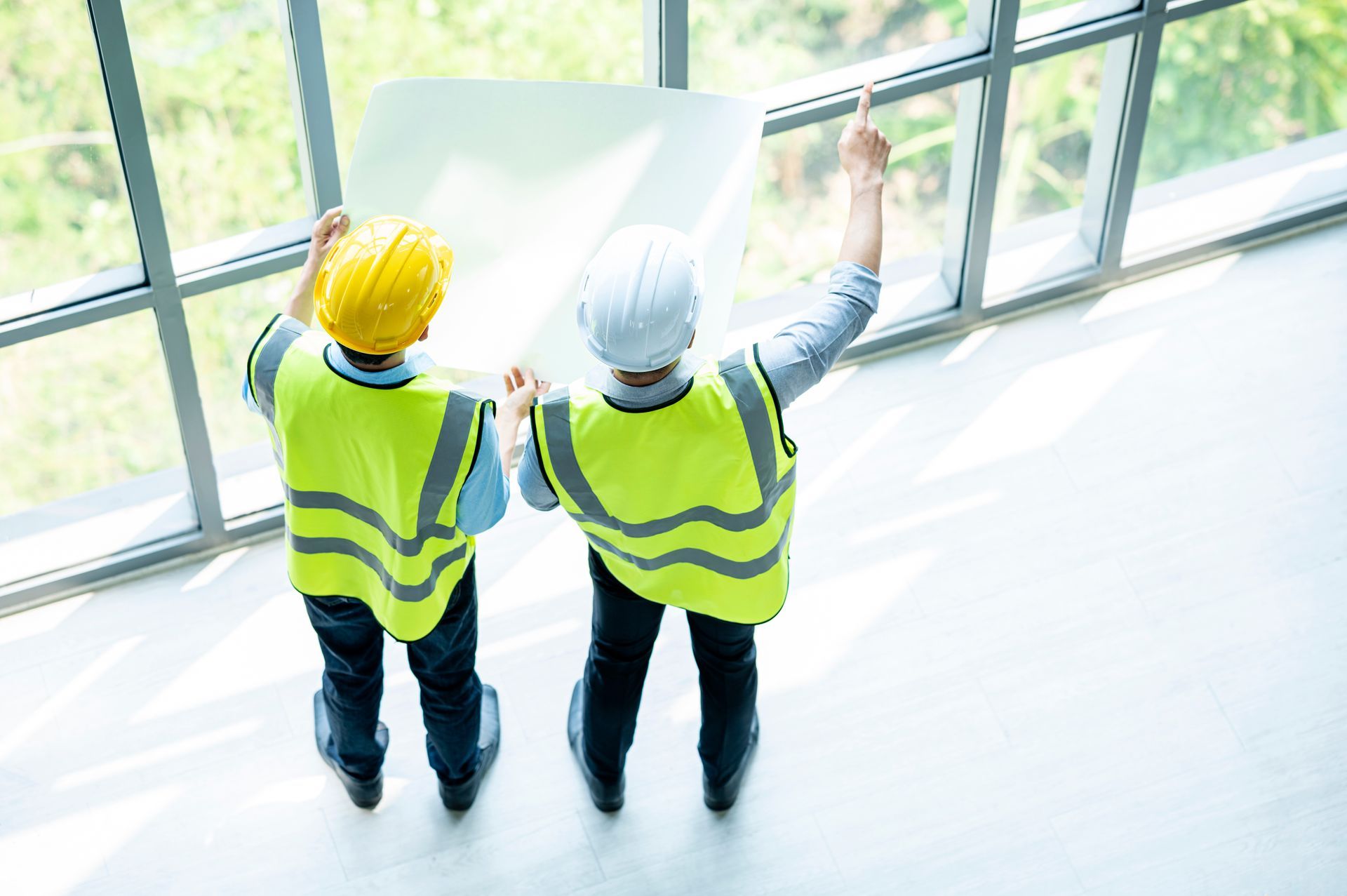 Due operai edili con giubbotti catarifrangenti e caschi protettivi esaminano i progetti vicino a una finestra.
