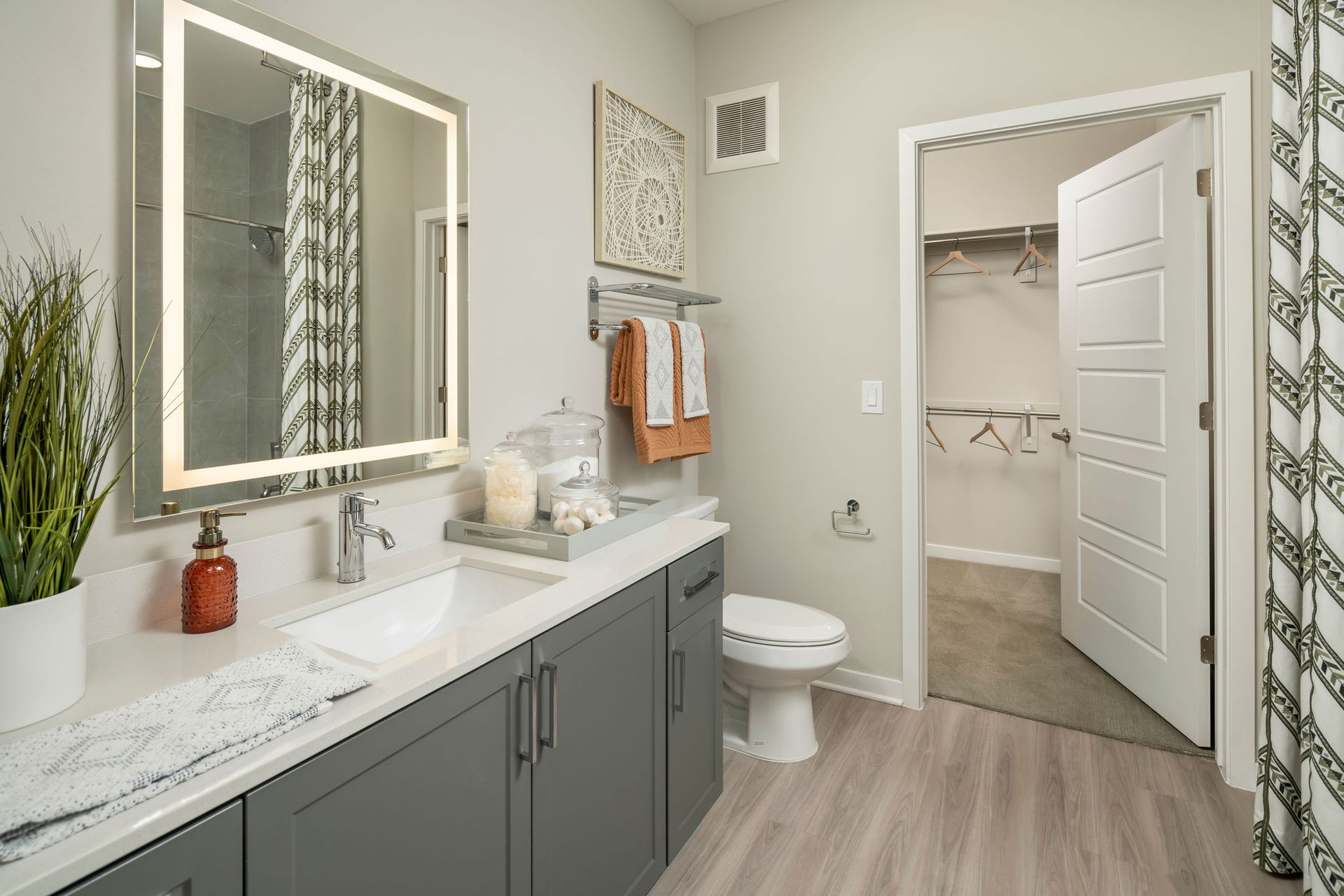 Modern bathroom with a gray vanity, backlit mirror, and a walk-in closet visible through the doorway.