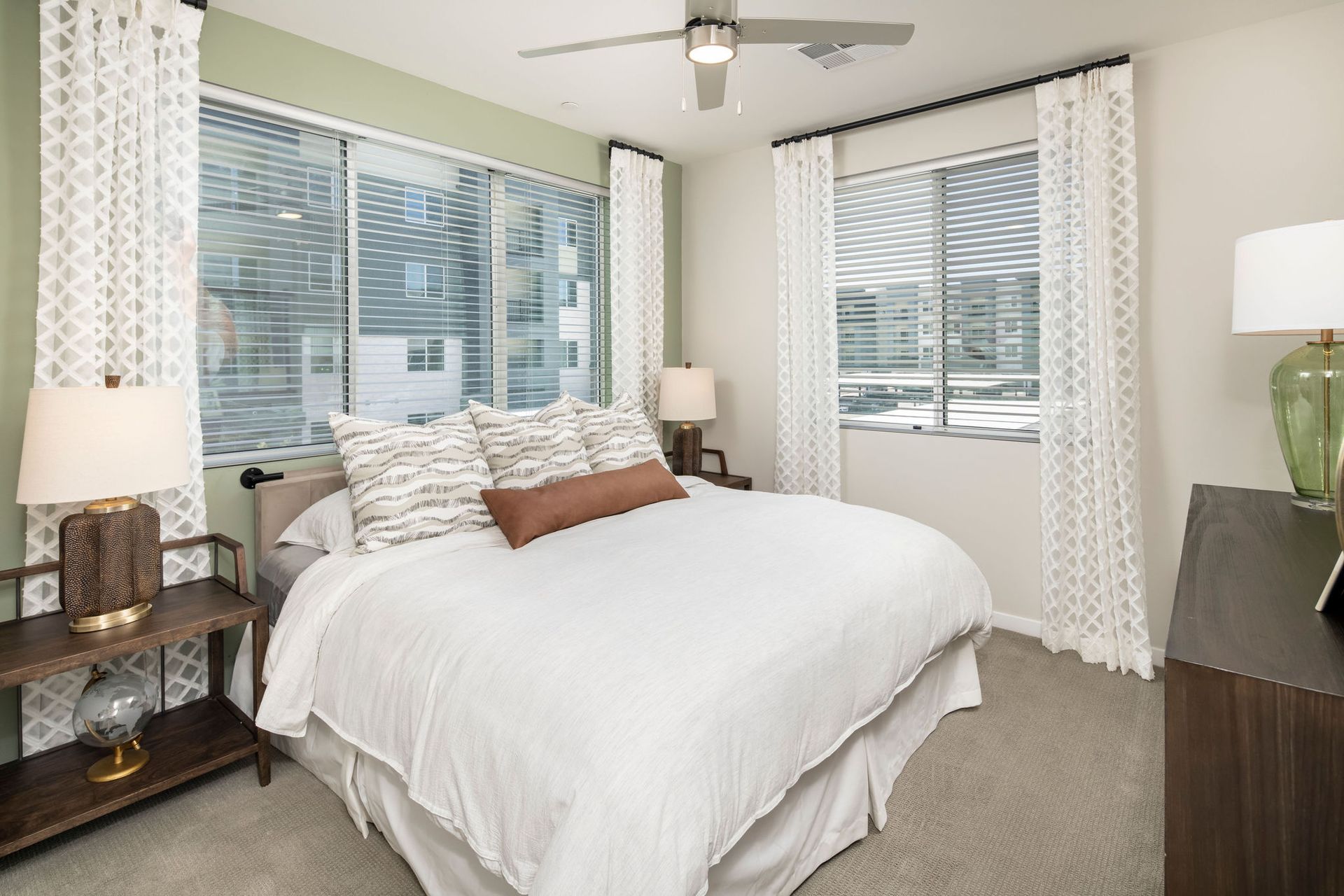 Furnished bedroom in an apartment with a large window, white curtains, and neutral decor.