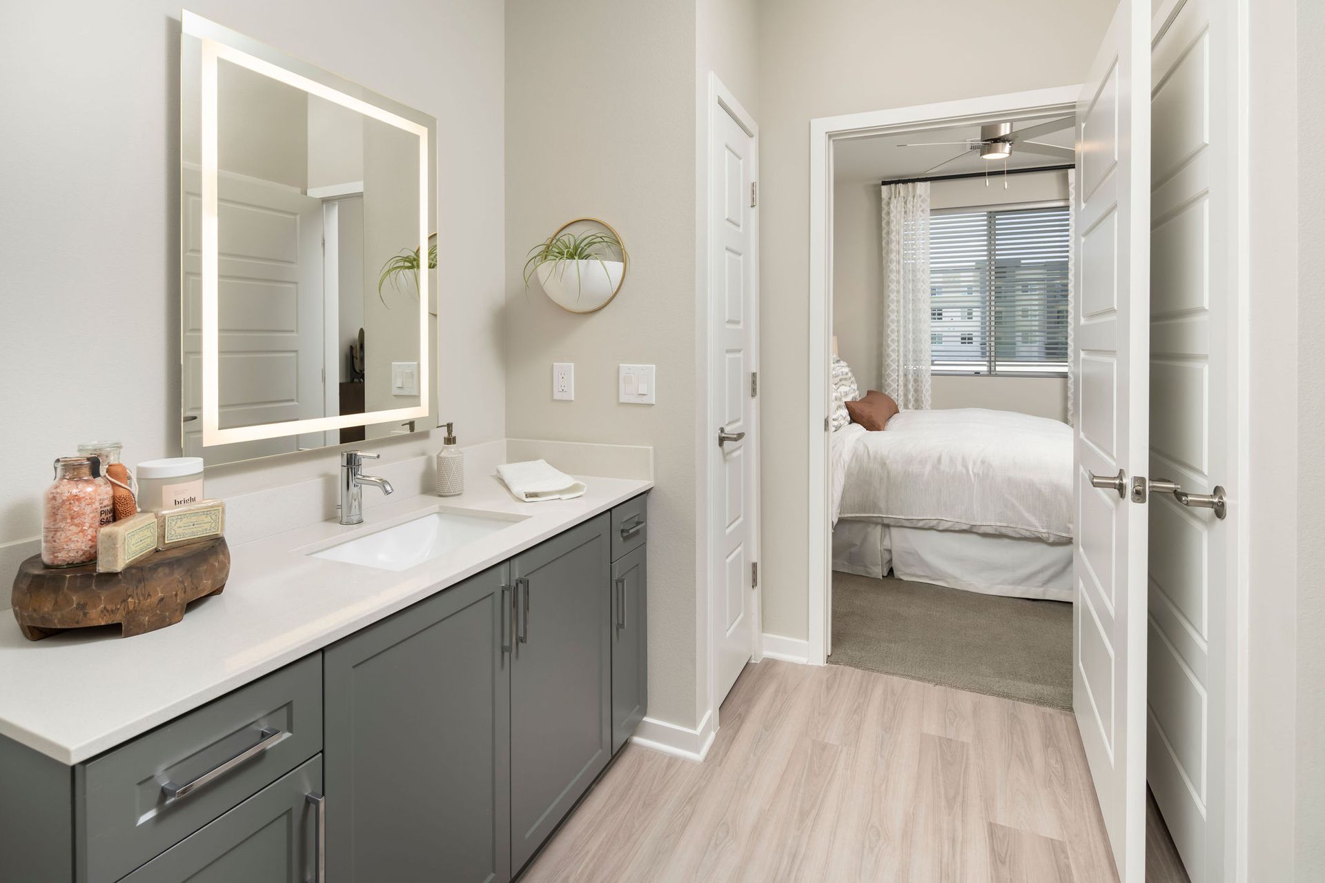 Bathroom vanity with gray cabinets, white countertop, backlit mirror, and doorway to a bedroom.