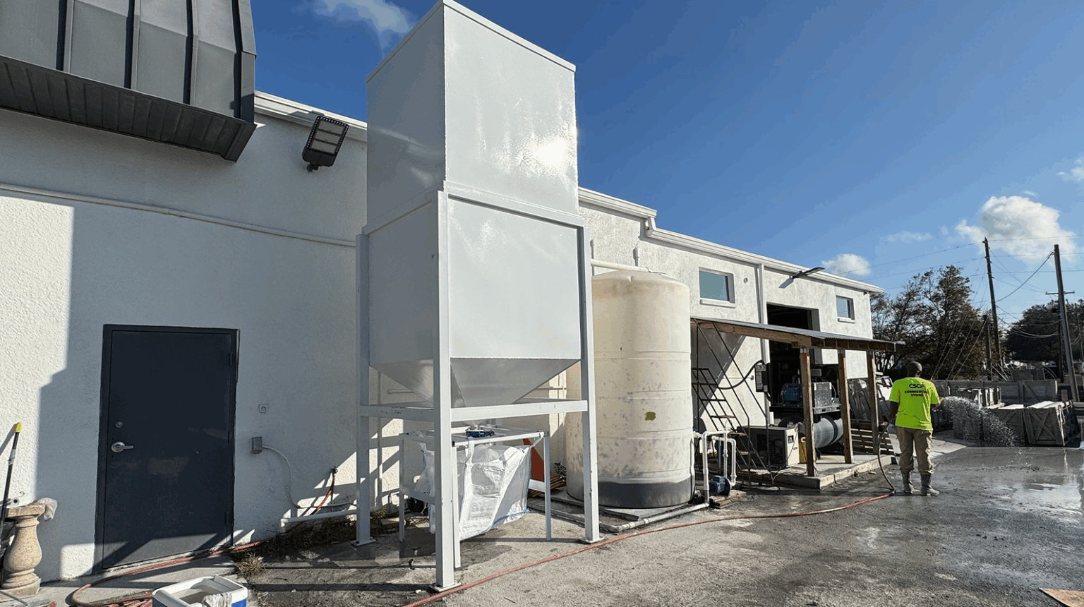White industrial filtration system with a tank, hopper, and bag against a white building and blue sky.