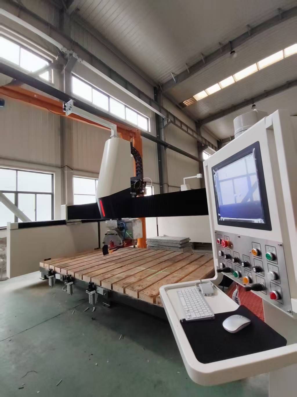 A large industrial CNC machine in a factory setting. It has a control panel, long arm, and wooden base.