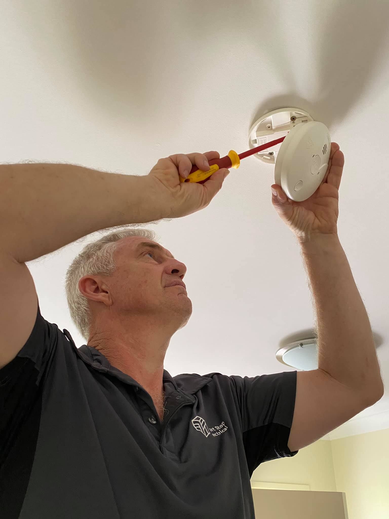 A man is fixing a smoke detector on the ceiling with a screwdriver. — Short Stop Electrical in Nerang, QLD