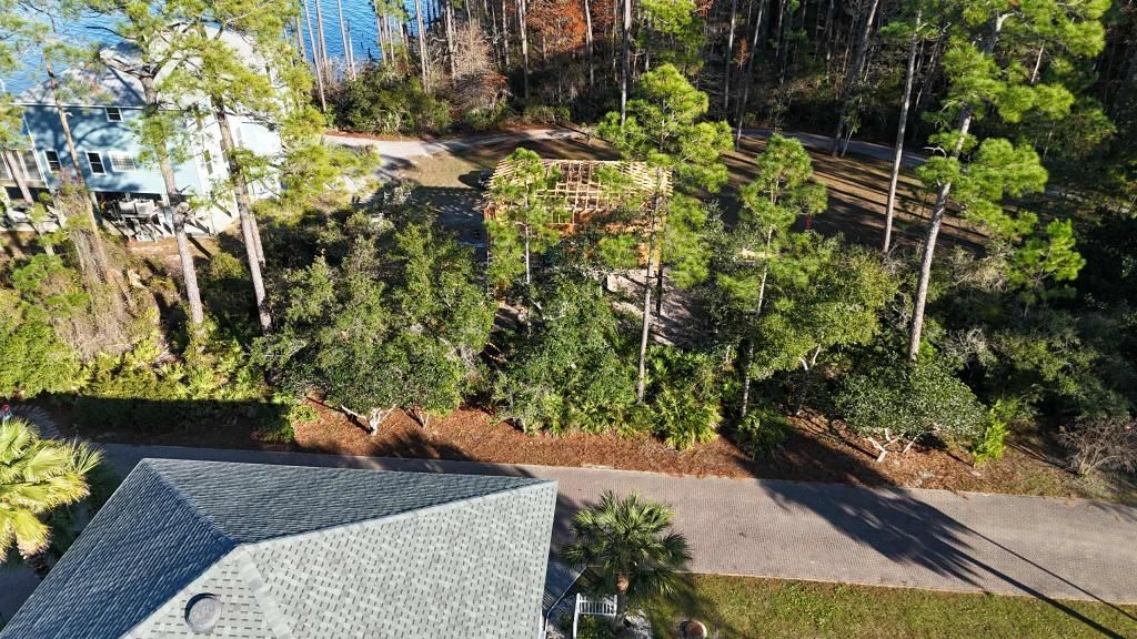 Overhead view of a house roof, road, and trees with a wooden structure in a clearing, next to a lake.