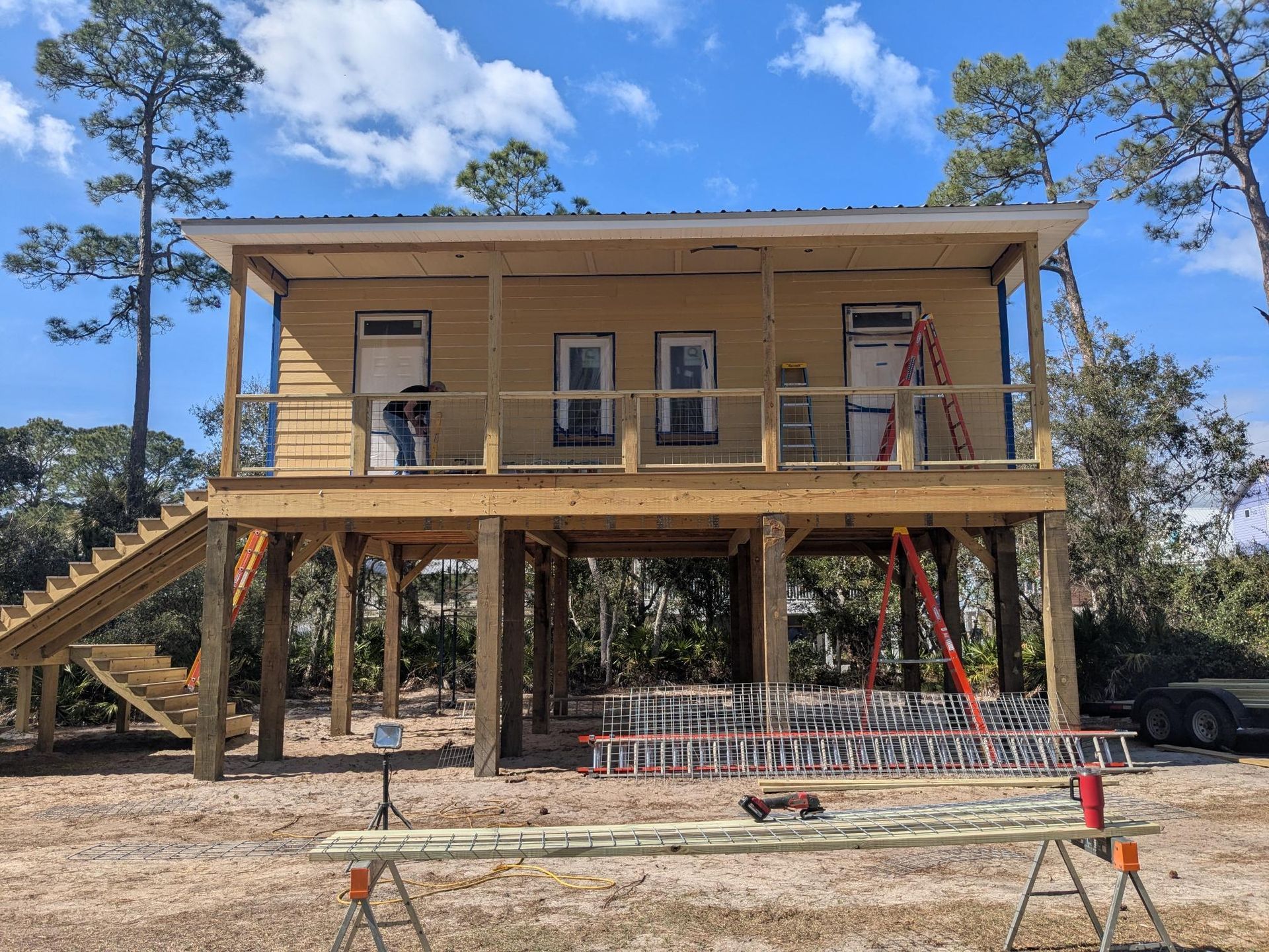 Elevated wooden house under construction; yellow siding, wrap-around porch, stairs, ladder, blue sky.