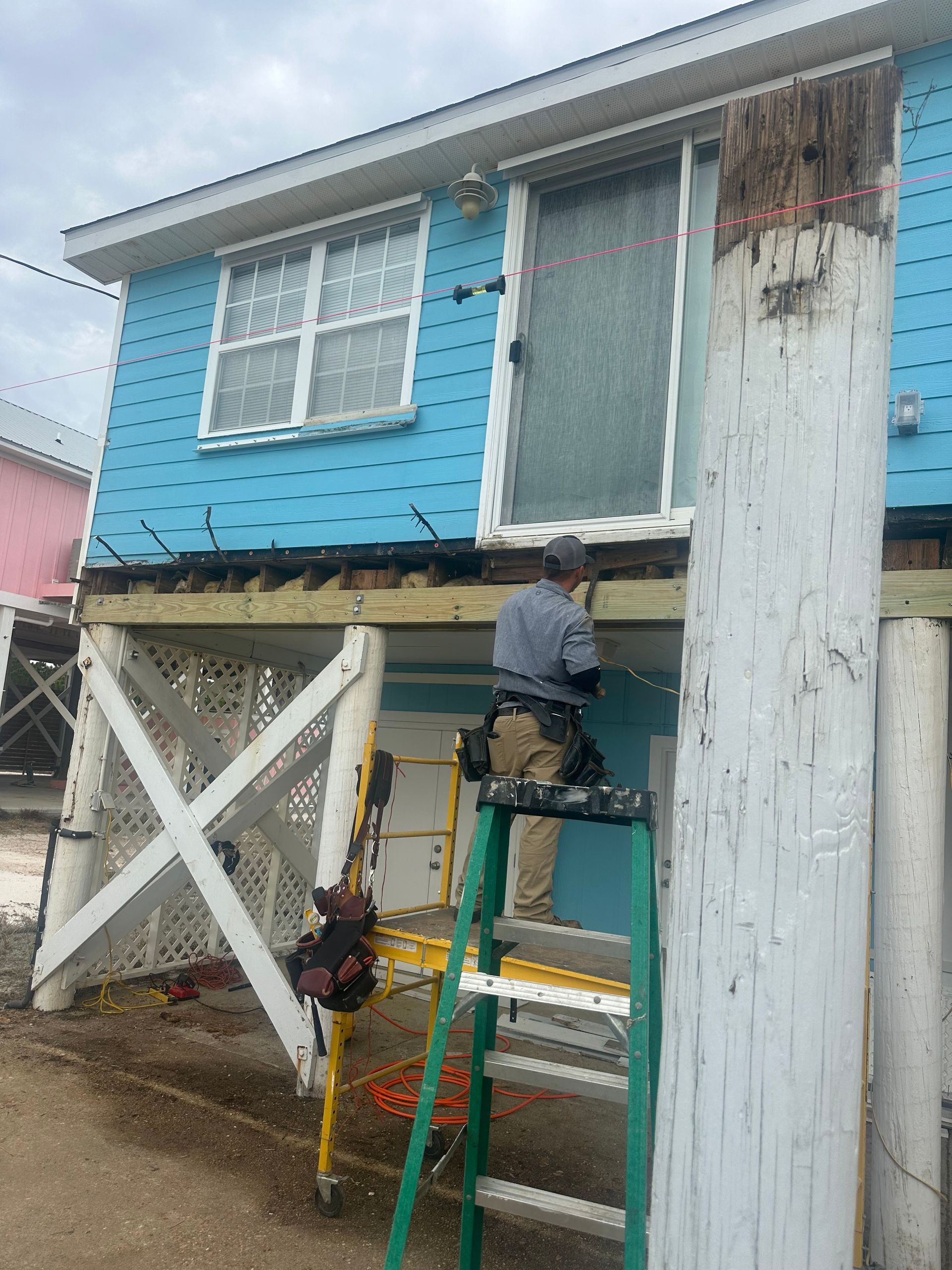 A person on a ladder repairs a blue house on stilts with white cross supports.