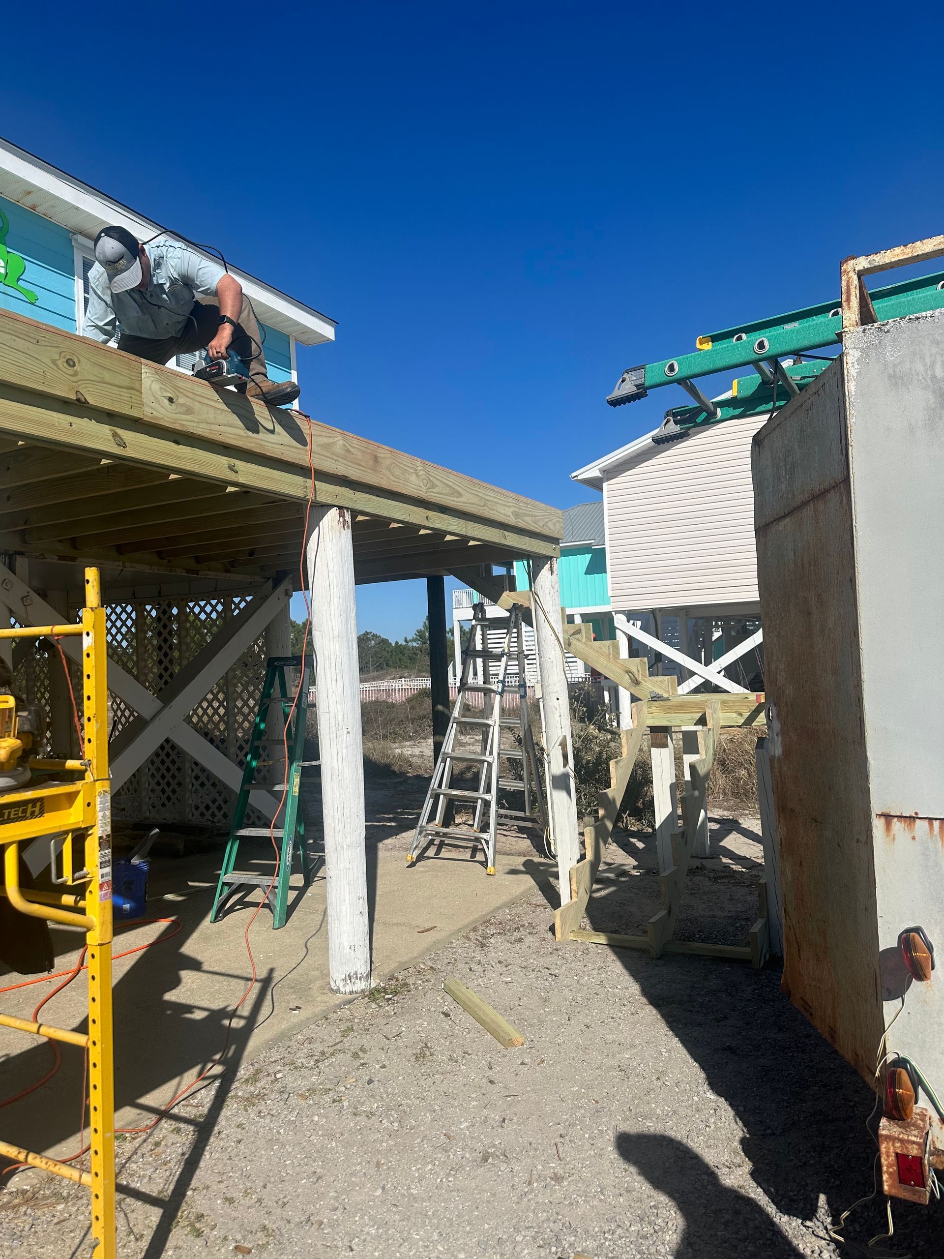 Construction worker on elevated deck, turquoise building, stairs, scaffolding, clear blue sky.