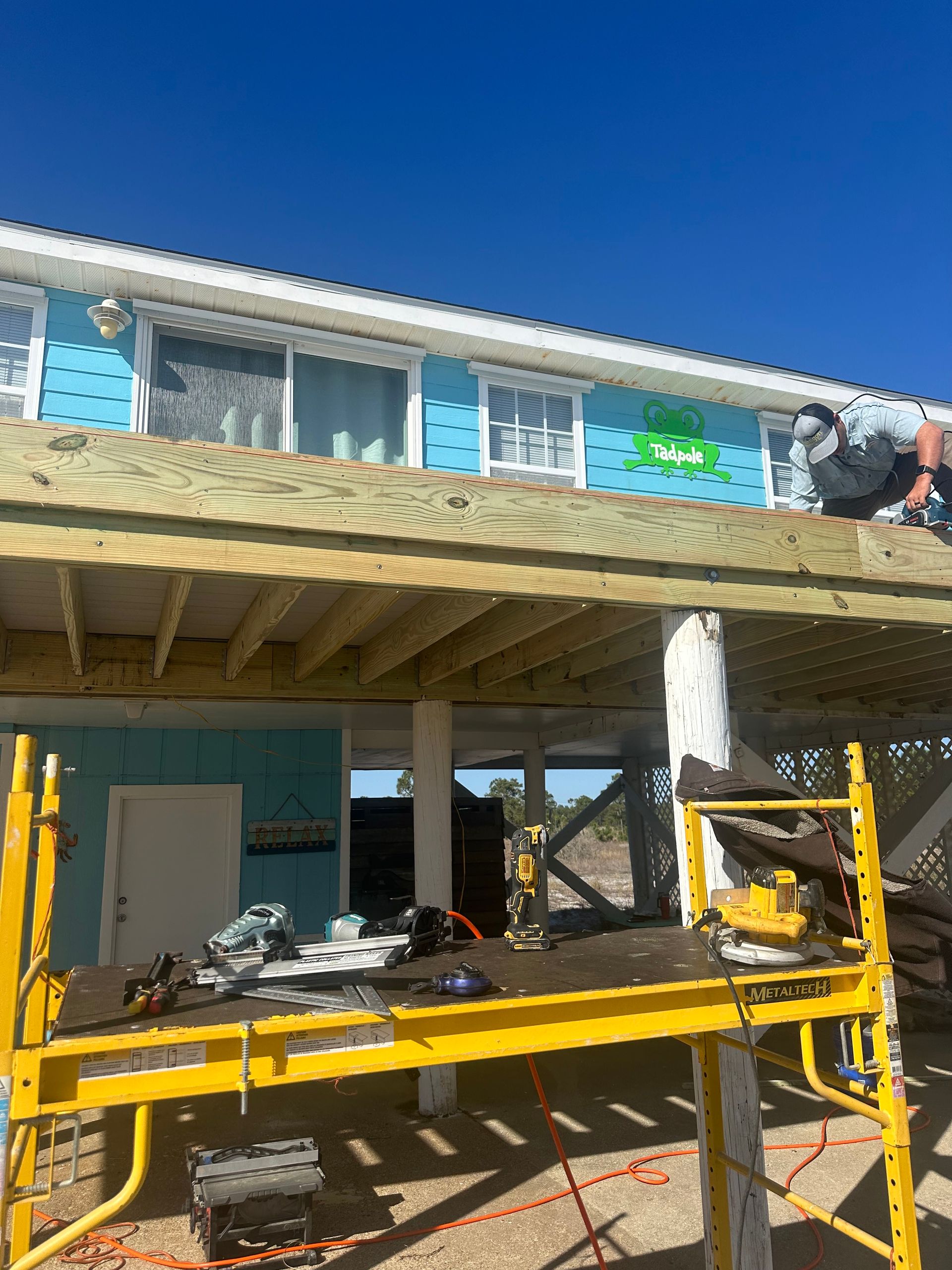 Construction on a deck; workers on the deck, scaffolding below; blue siding, clear sky.
