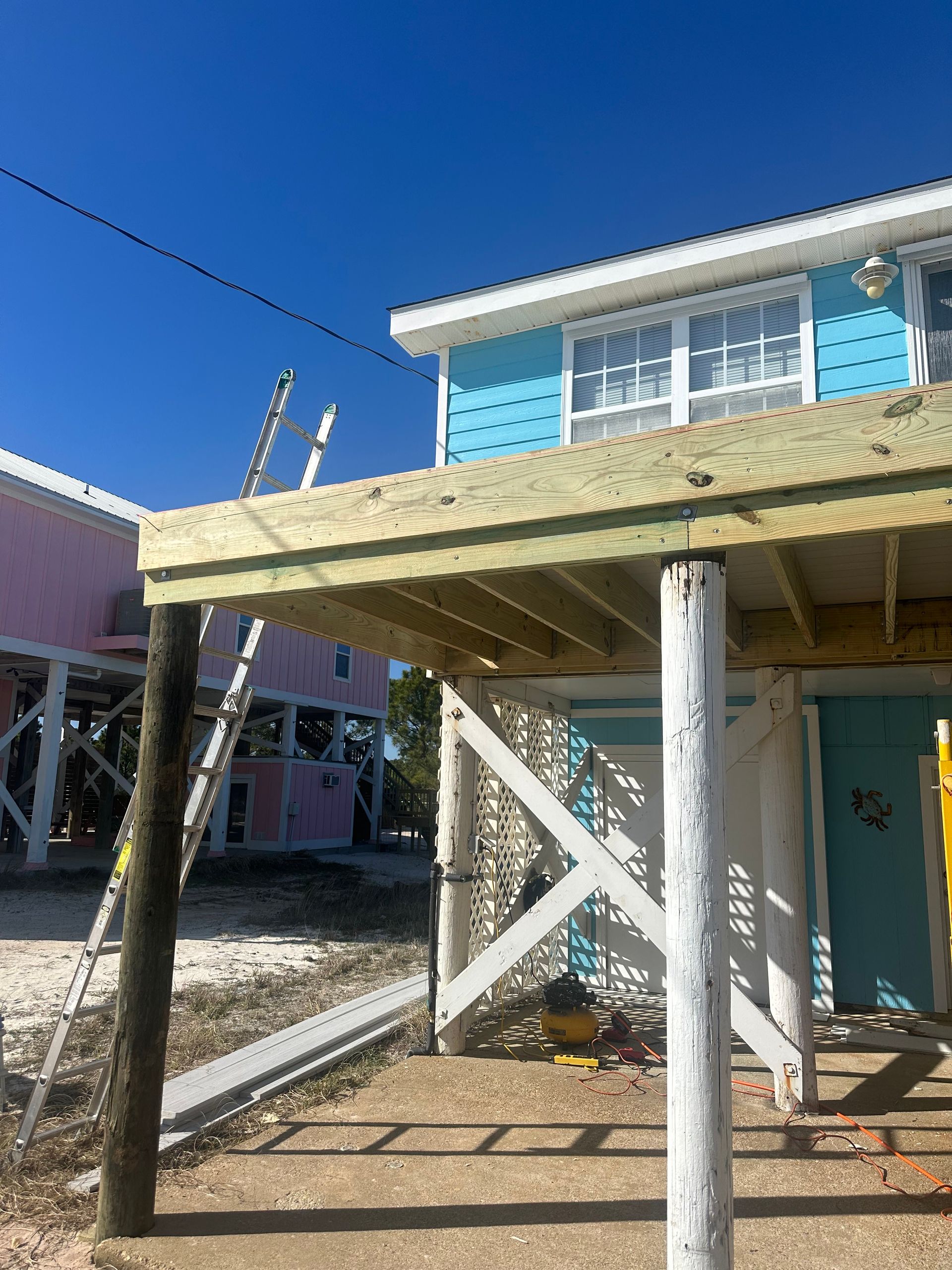 A two-story blue beach house with an elevated deck under construction; pink house in the background.