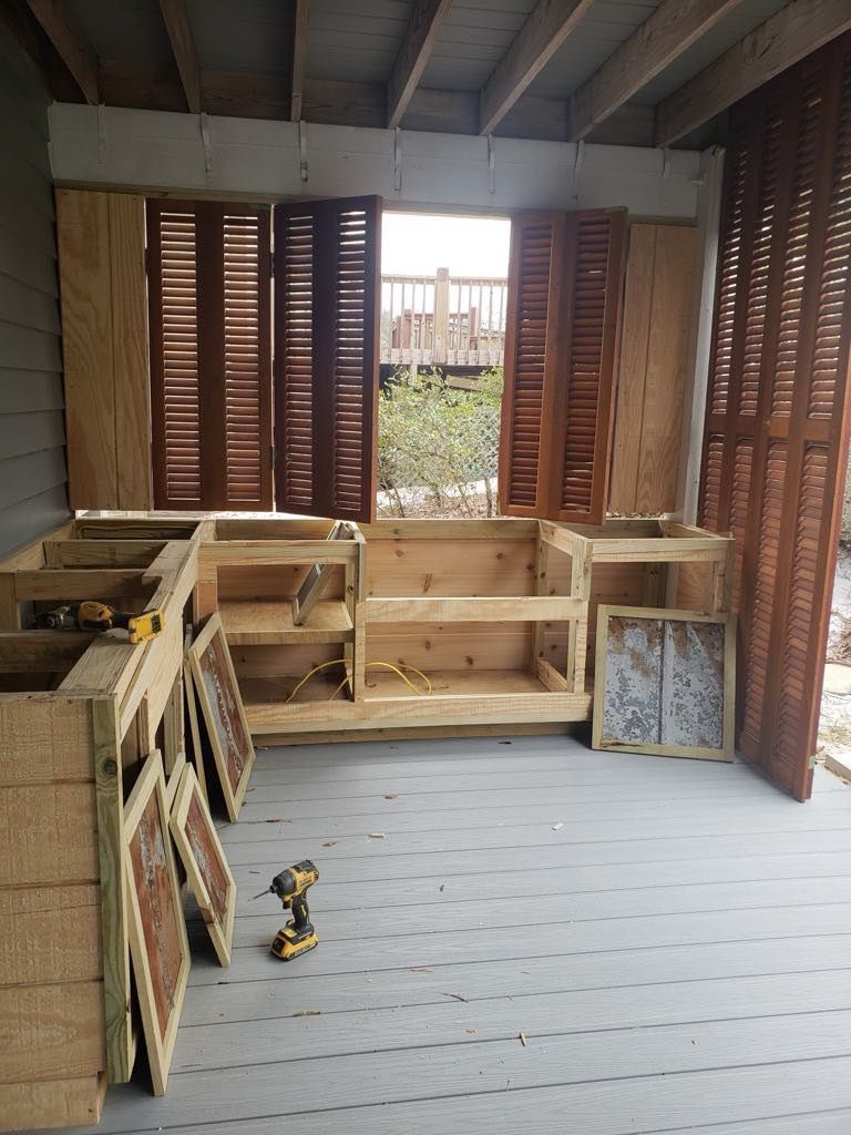 Wooden outdoor kitchen under construction; shutters, shelves, and cabinets. Gray deck.