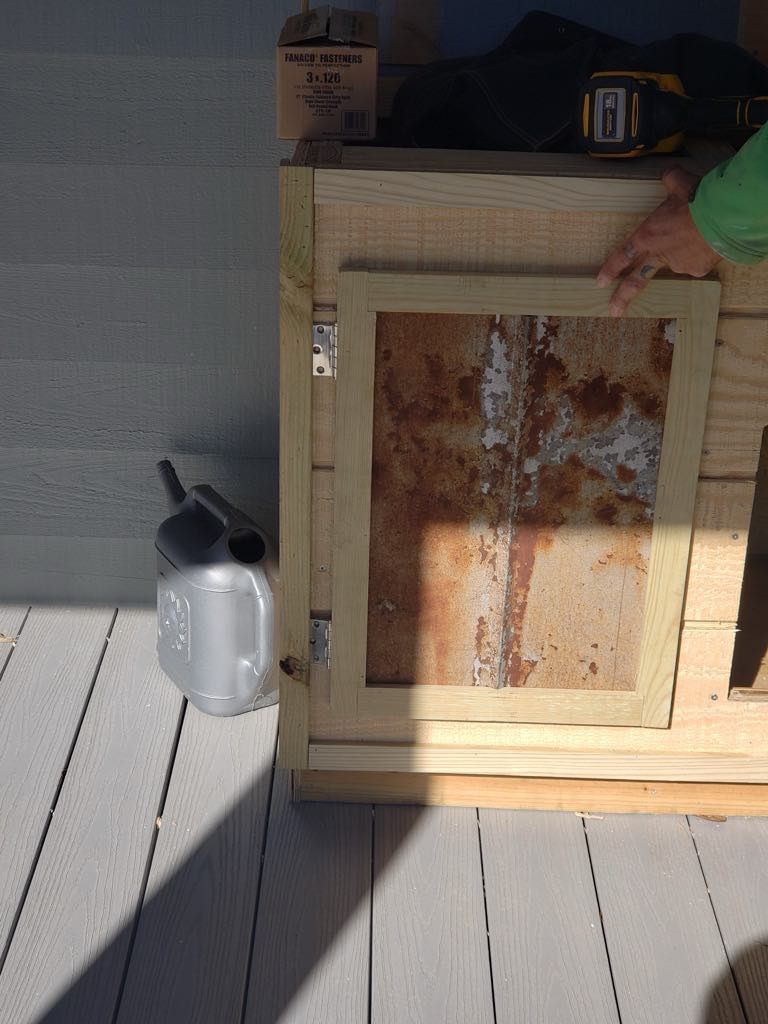 Wooden box with a rusty metal door on a deck, with a watering can nearby.