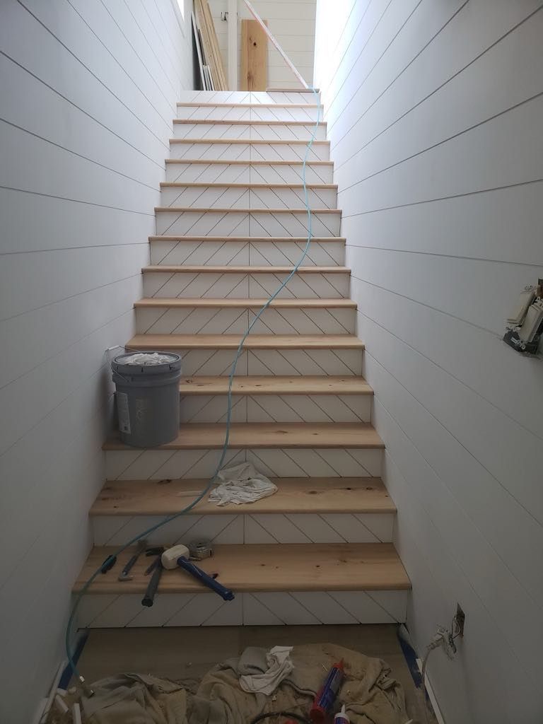 Staircase under construction: unfinished wooden steps with angled patterns, surrounded by white shiplap walls.