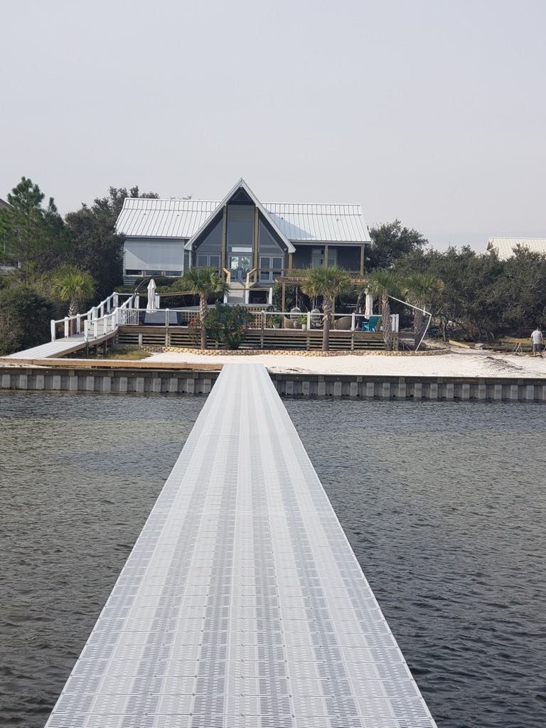 A house with a pier extending over the water. White pier leads to a light blue house with a white roof.