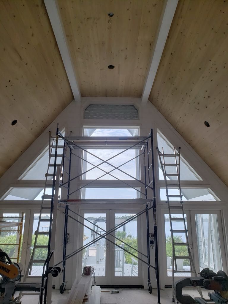Interior of a room under construction with scaffolding and large windows.  Light wood ceiling and white trim.