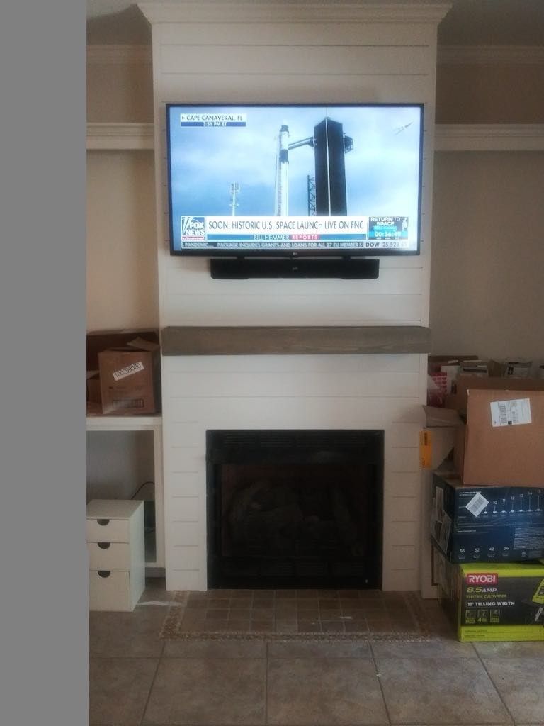 TV above fireplace showing a news broadcast. Fireplace is flanked by shelves and construction items.