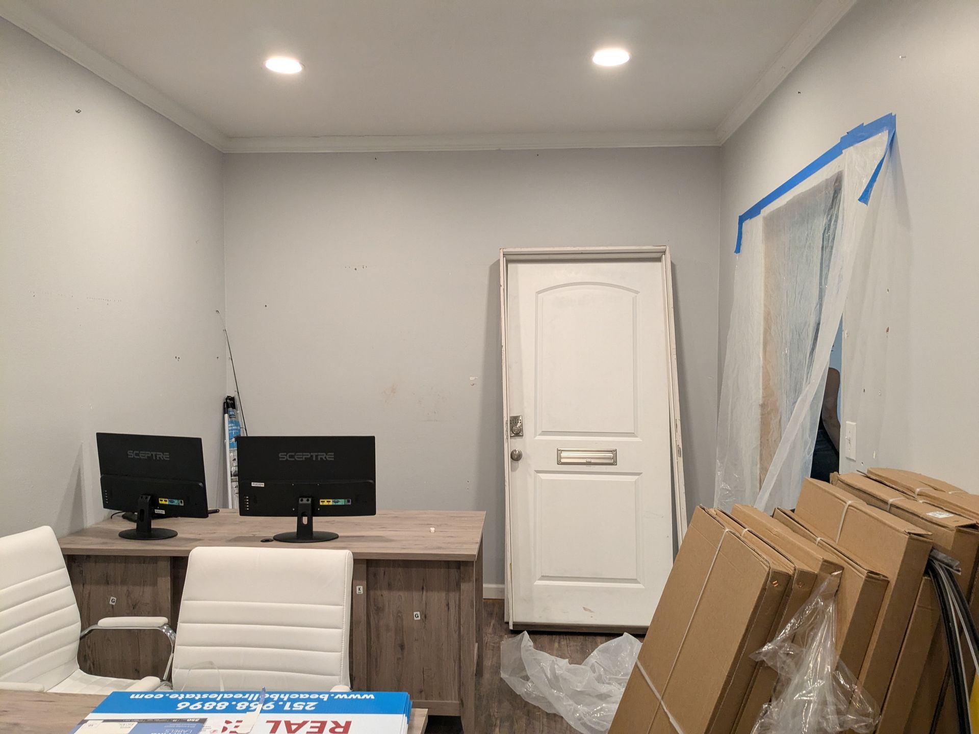 Office with desk, computers, and door, under construction with plastic sheeting and cardboard boxes.
