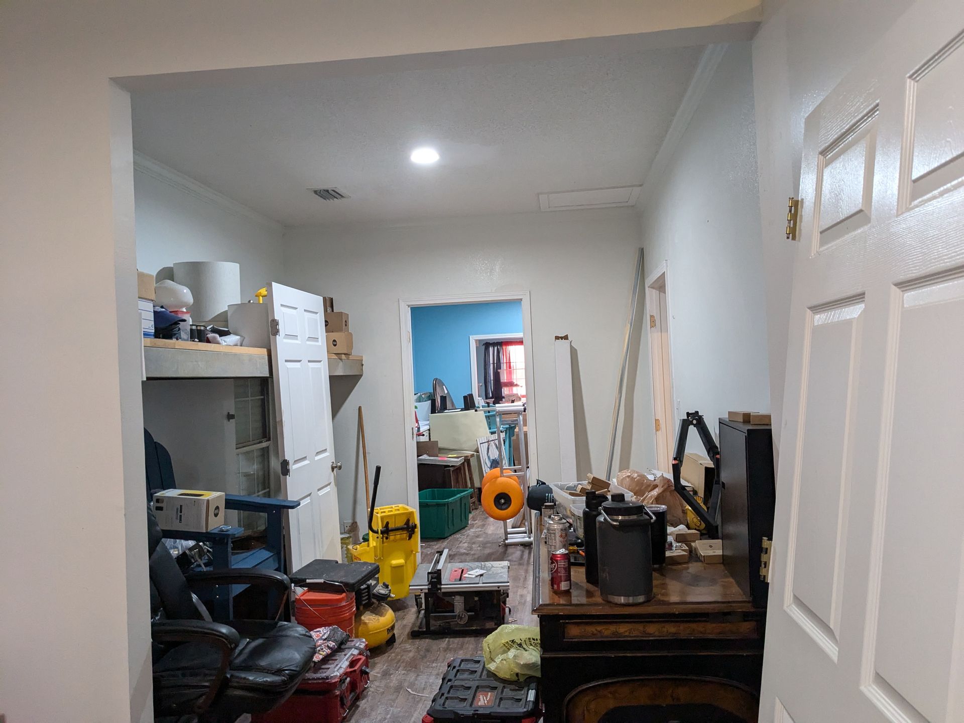 Cluttered room: tools, furniture, open doorways. White walls, wood floor.