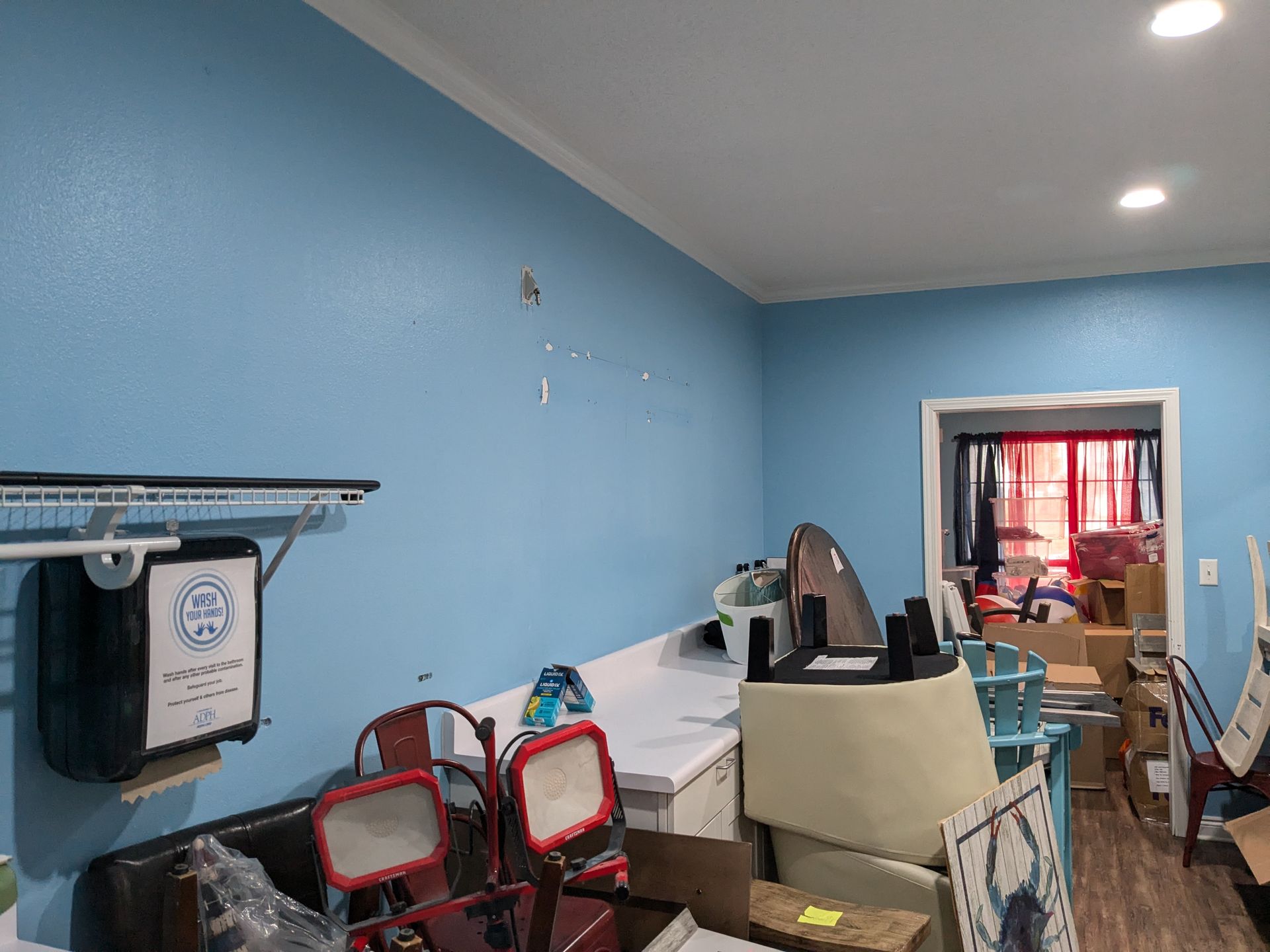 Blue-painted room with clutter; laundry area with ironing board and appliances; open doorway into another room.