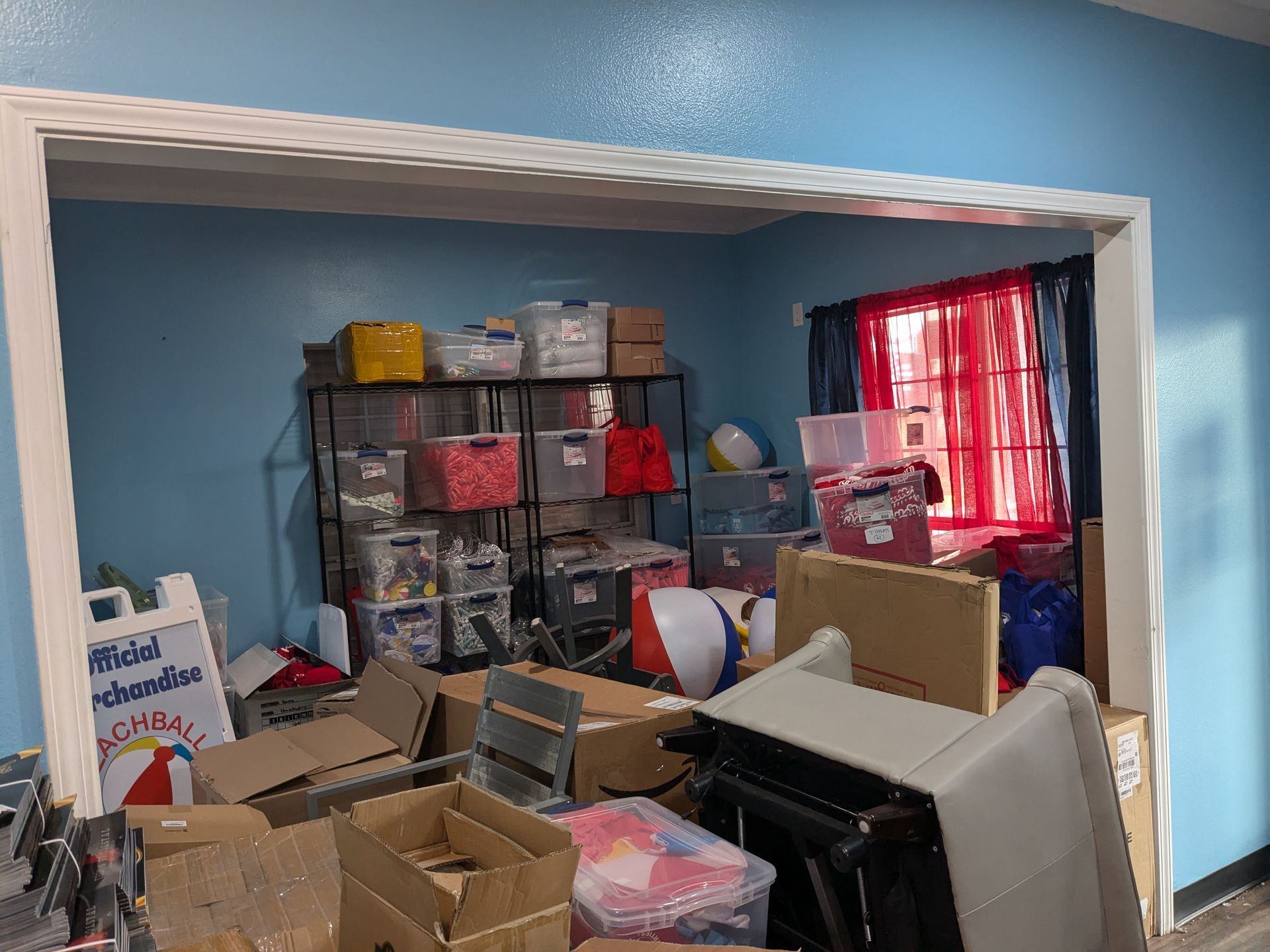 Room with blue walls, cluttered with boxes, storage bins, and a metal shelf. Red curtained window.
