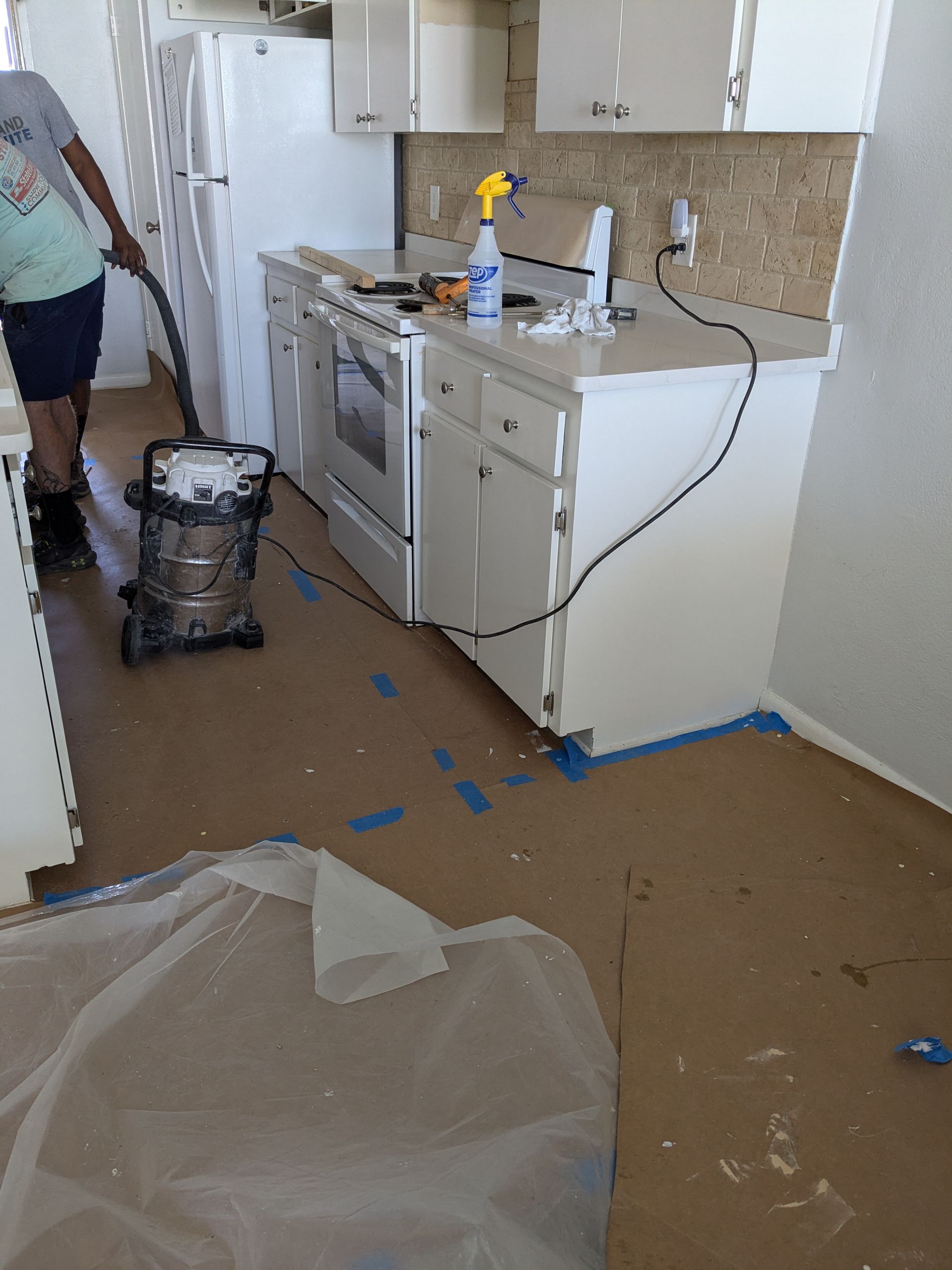 Person vacuuming a kitchen with white cabinets, appliances, and tan floor.