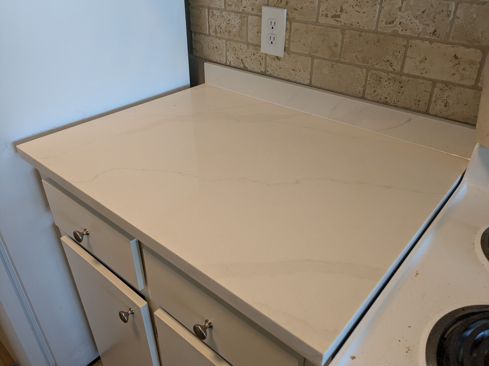 White countertop on kitchen cabinets, next to a stove and against a brick wall.