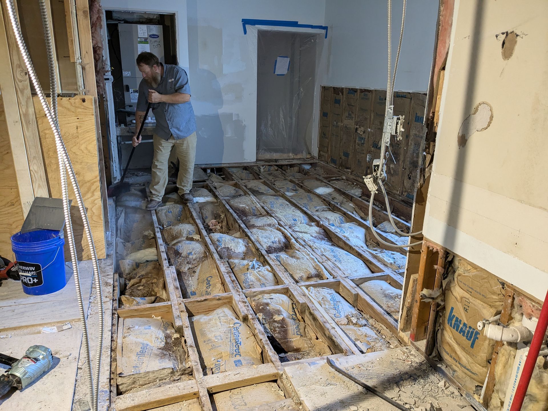Man removing flooring in a room during renovation. Exposed joists, debris, and tools are visible.