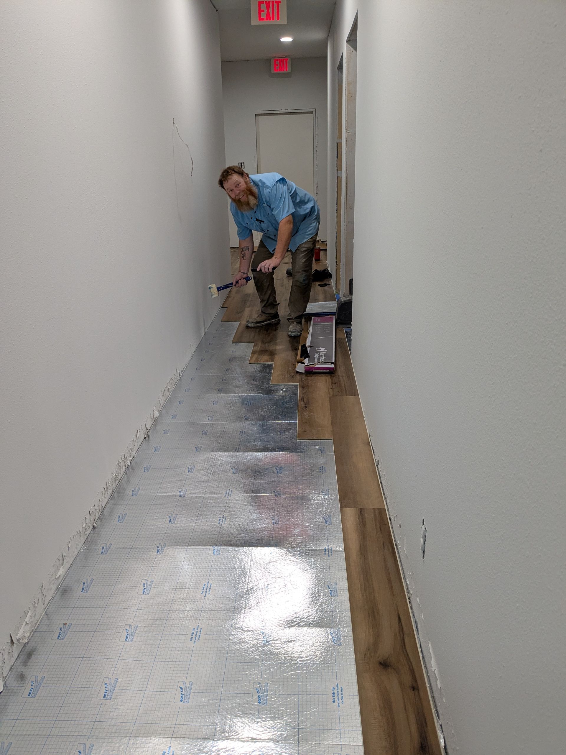 Person installing flooring in a narrow hallway. They are using a tool on wet, shiny flooring. White walls, wood-look planks.