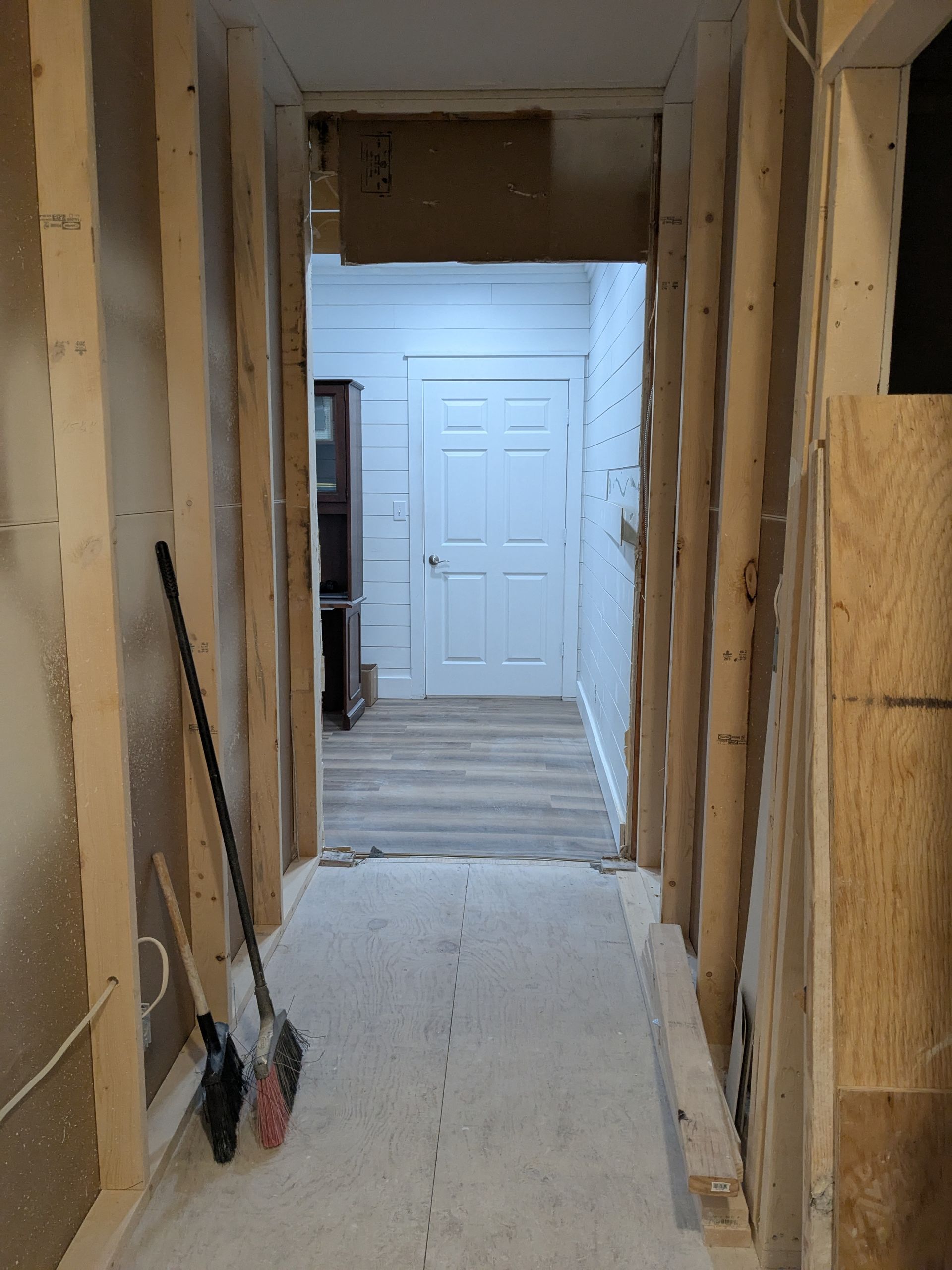 Hallway under construction: wood framing, doorway at end leads to a white door, tools lean against studs.