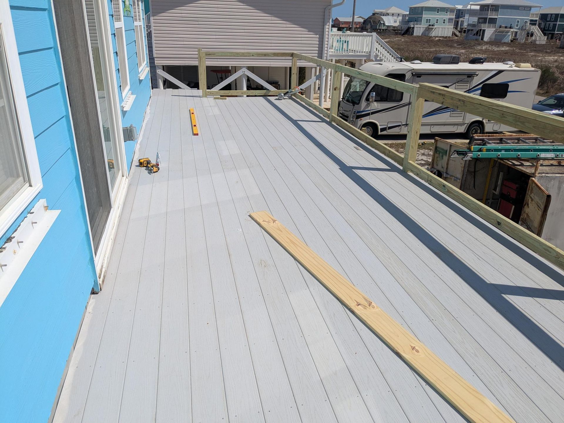 A gray wooden deck with unfinished railing on a blue house. RV and vehicles in background.