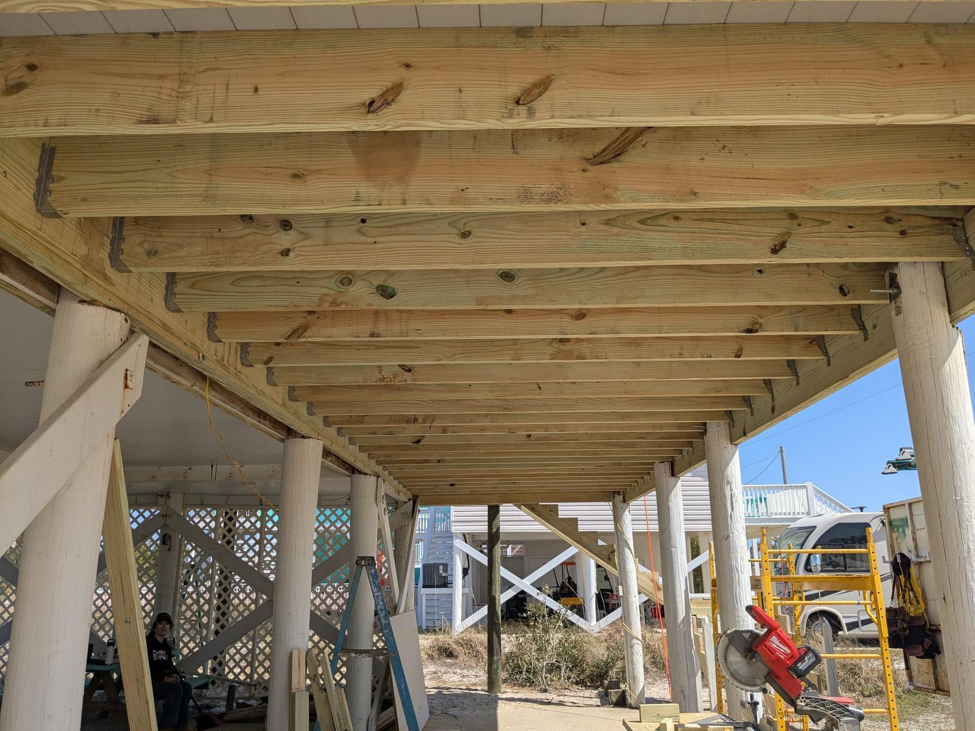Underside of a wooden deck under construction, supported by white columns. A person stands nearby.