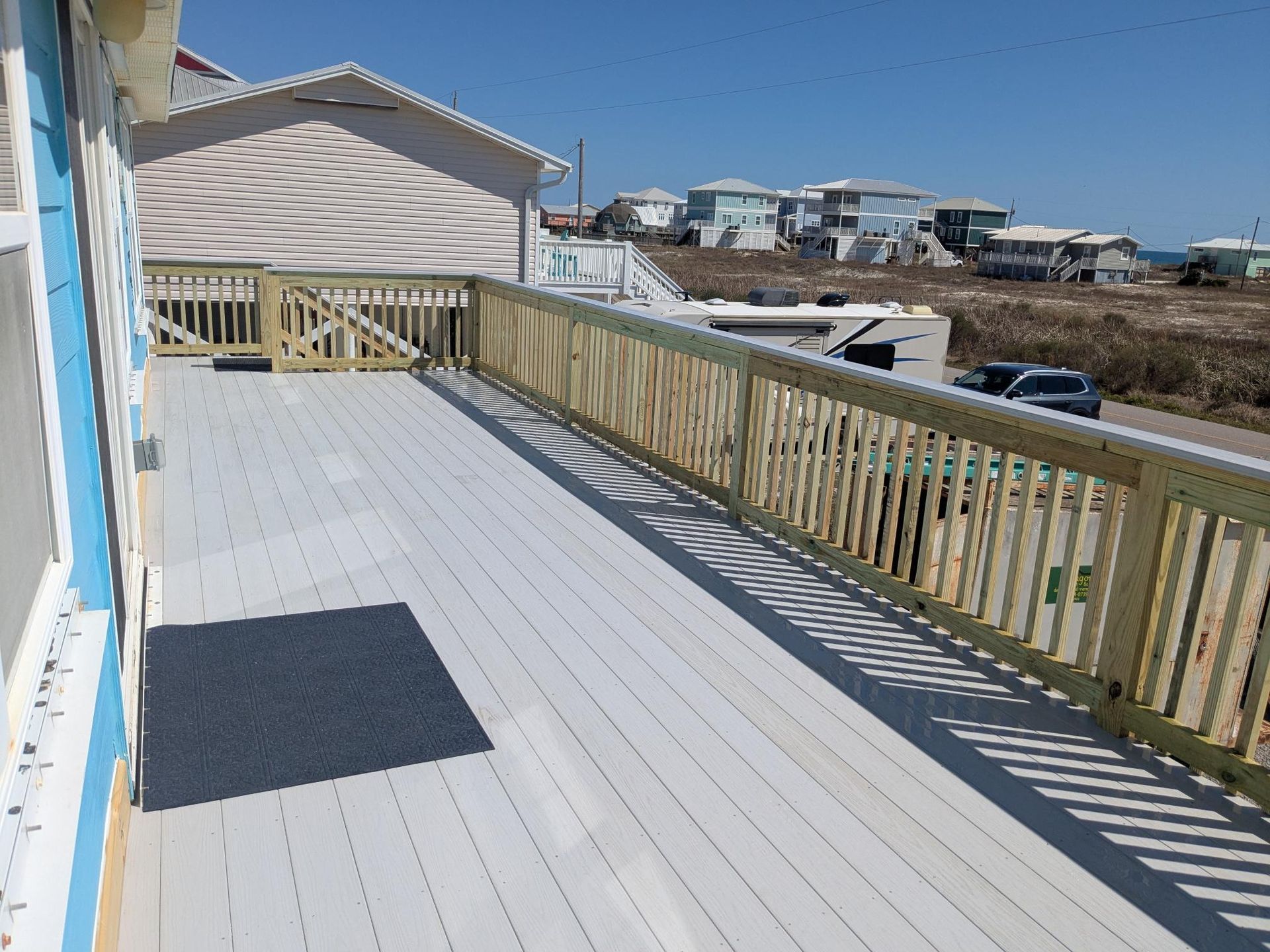 Wooden deck with railing, house in the background. Blue sky, ocean view.