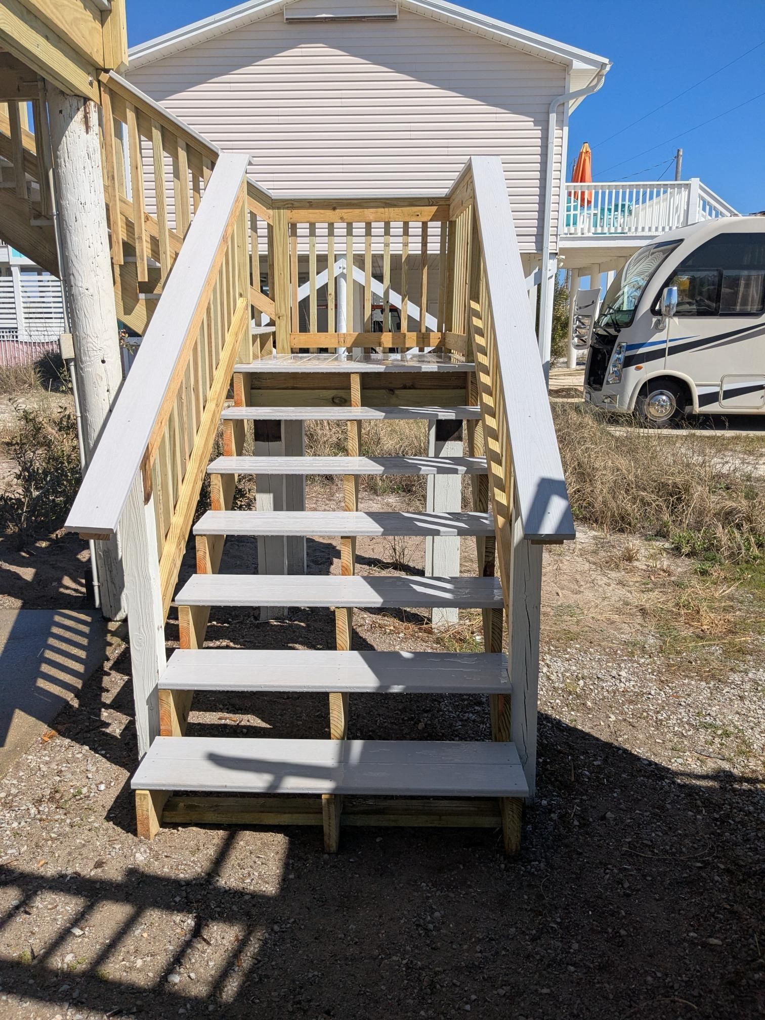 Wooden outdoor stairs leading up to a raised white beach house. Gray steps, beige railings, RV in background.