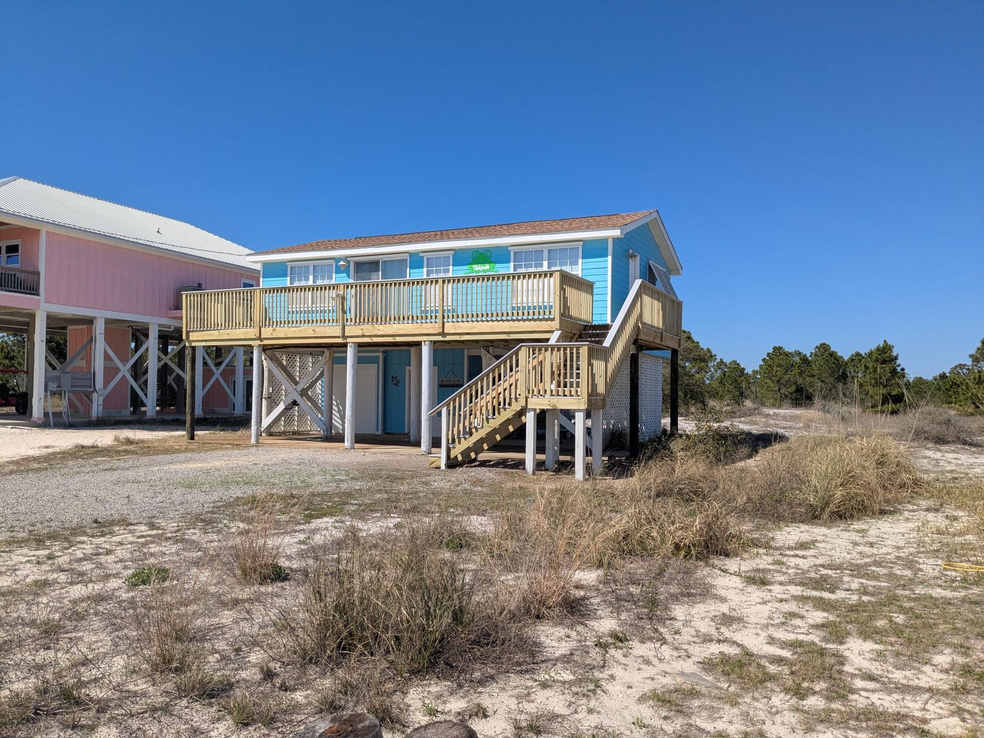 Blue beach house with deck, wooden stairs, and surrounding scrub brush under a clear blue sky.
