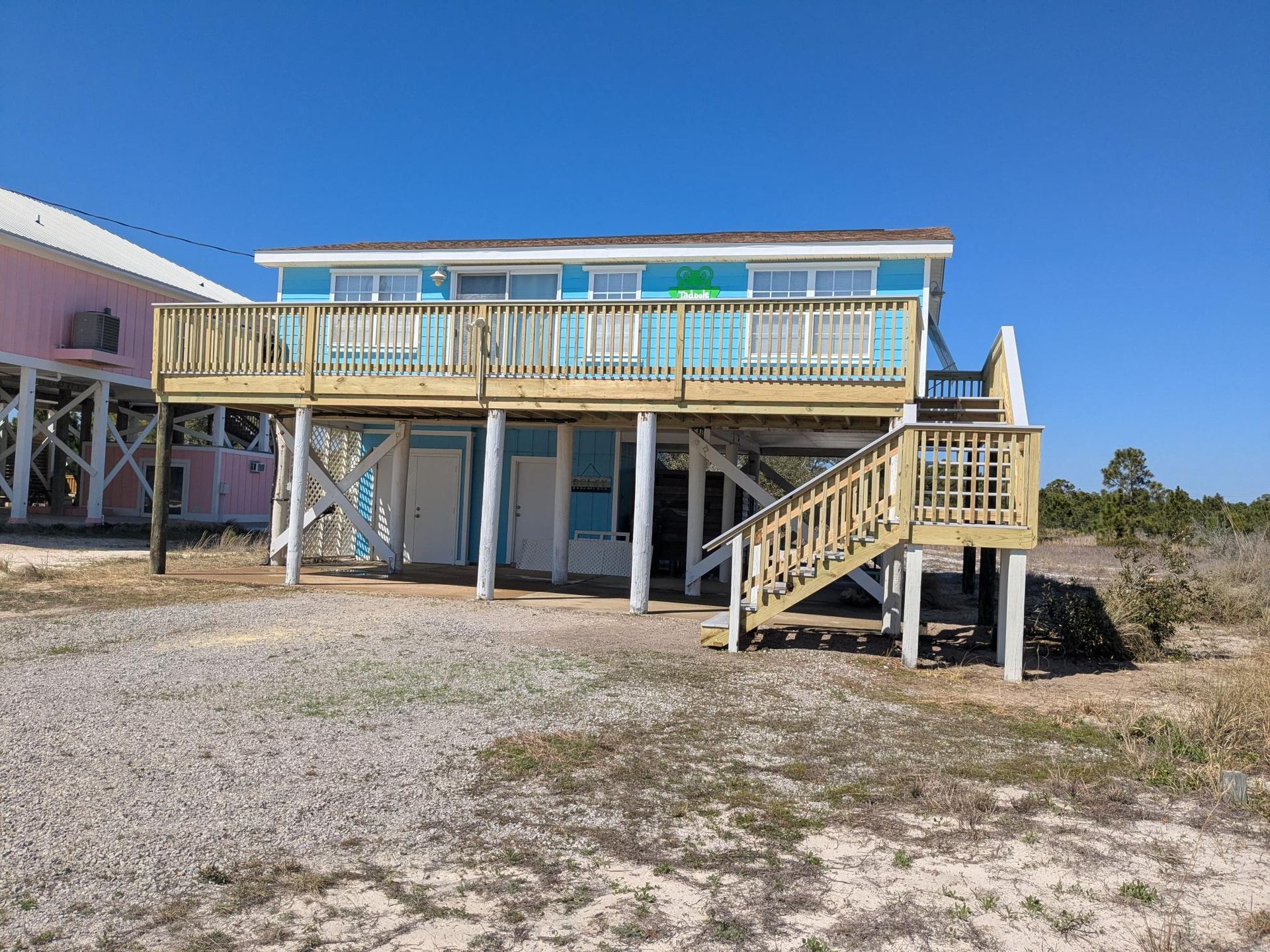 Two-story beach house on stilts with blue siding, a wooden deck, and a pink house next door.