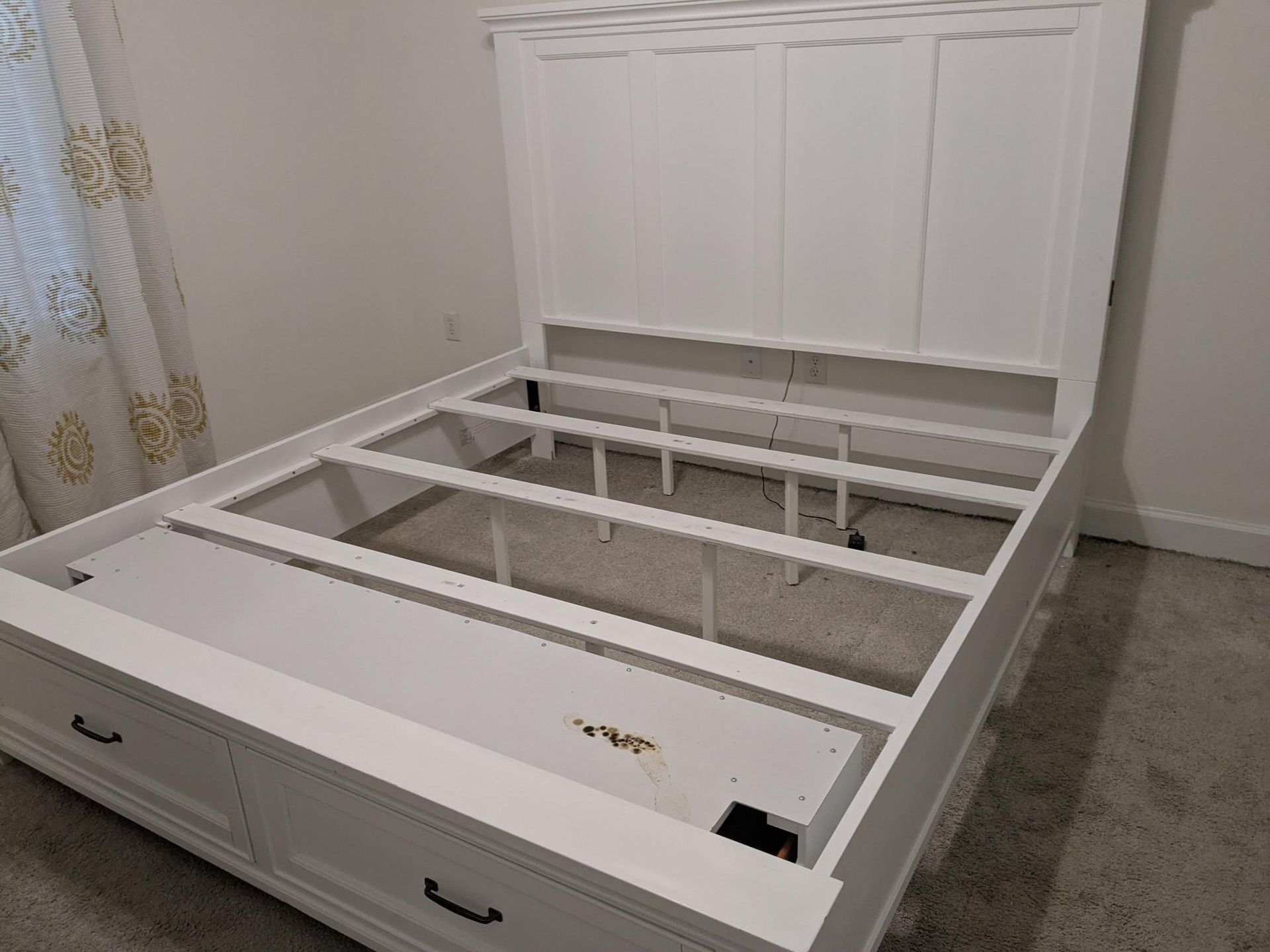White bed frame with headboard and storage drawers, in a bedroom with gray carpet.
