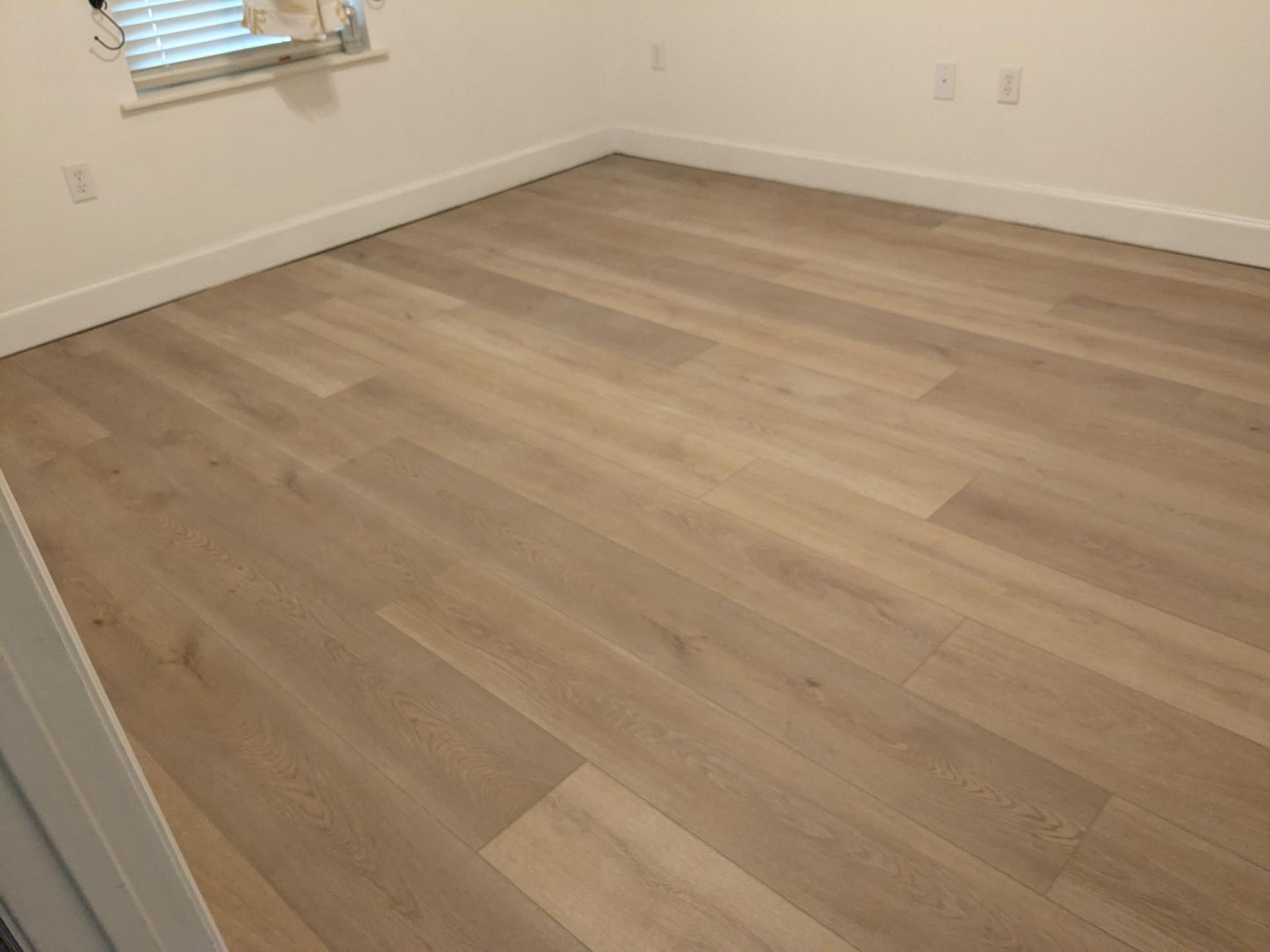 Light wood-look flooring in a room with white baseboards and walls. A window with blinds is on the left.