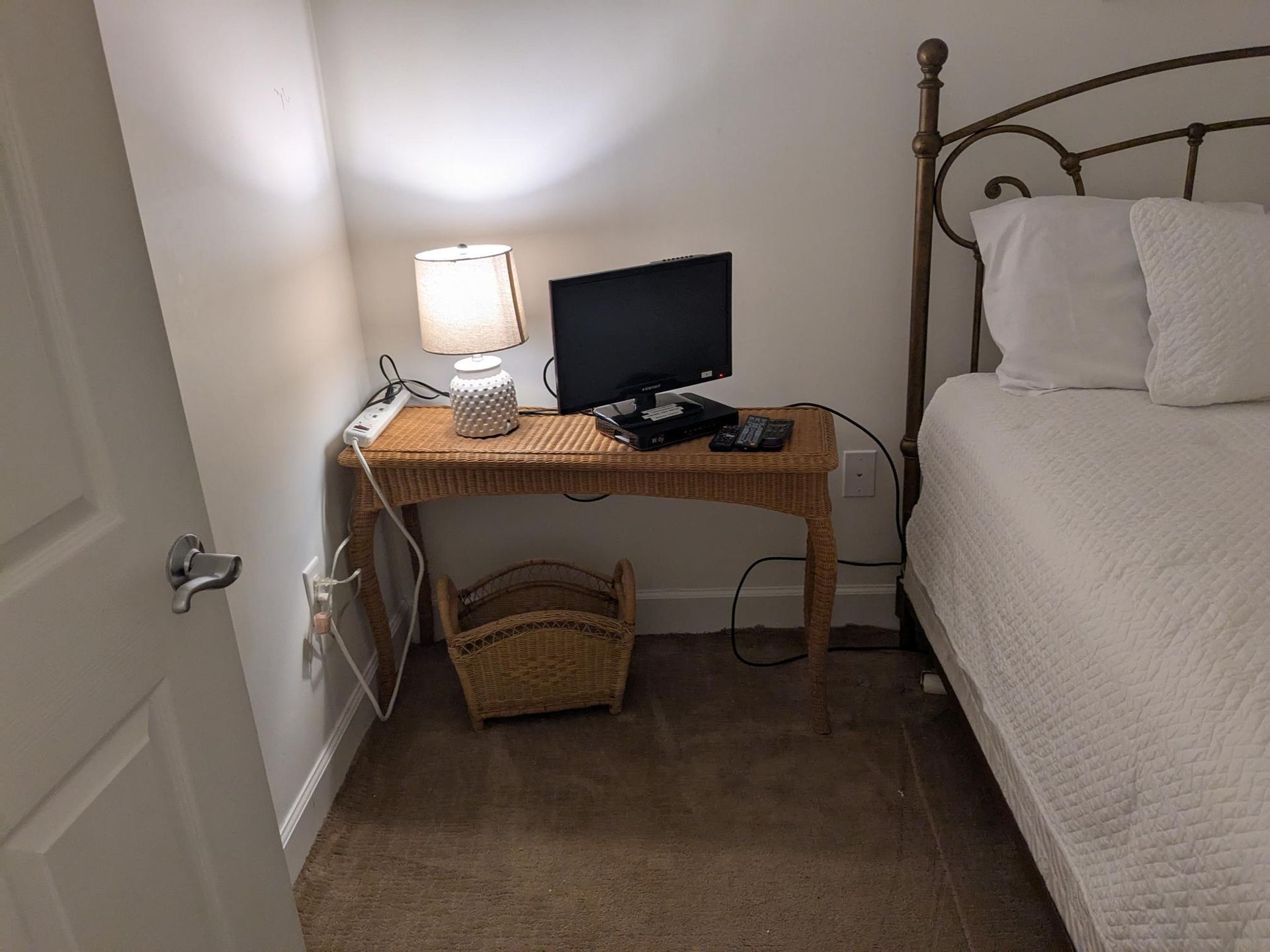 A bedroom with a small desk holding a lamp, monitor, and basket. A bed is next to the desk.