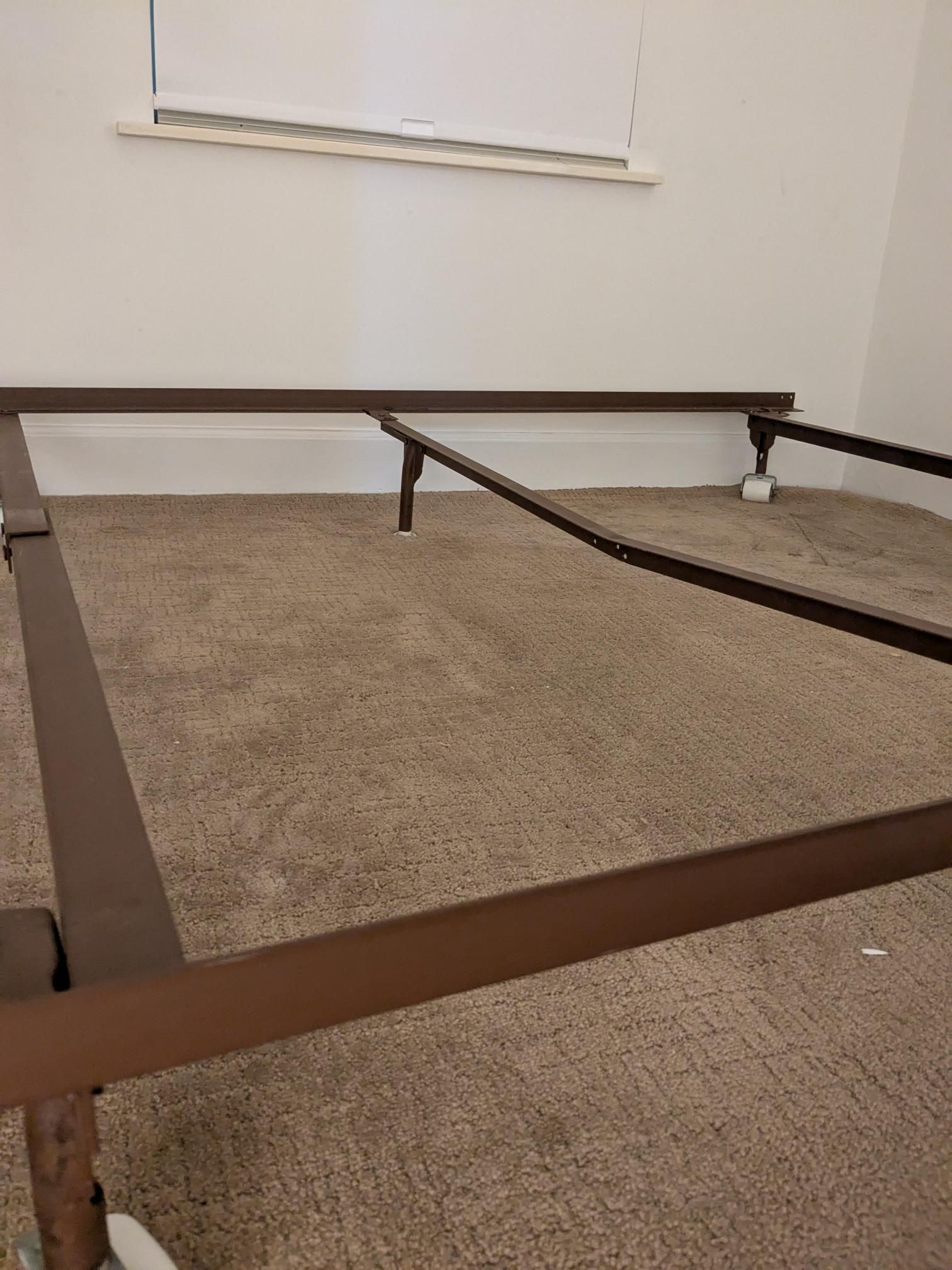 Brown metal bed frame on a carpeted floor, against a white wall with a window.
