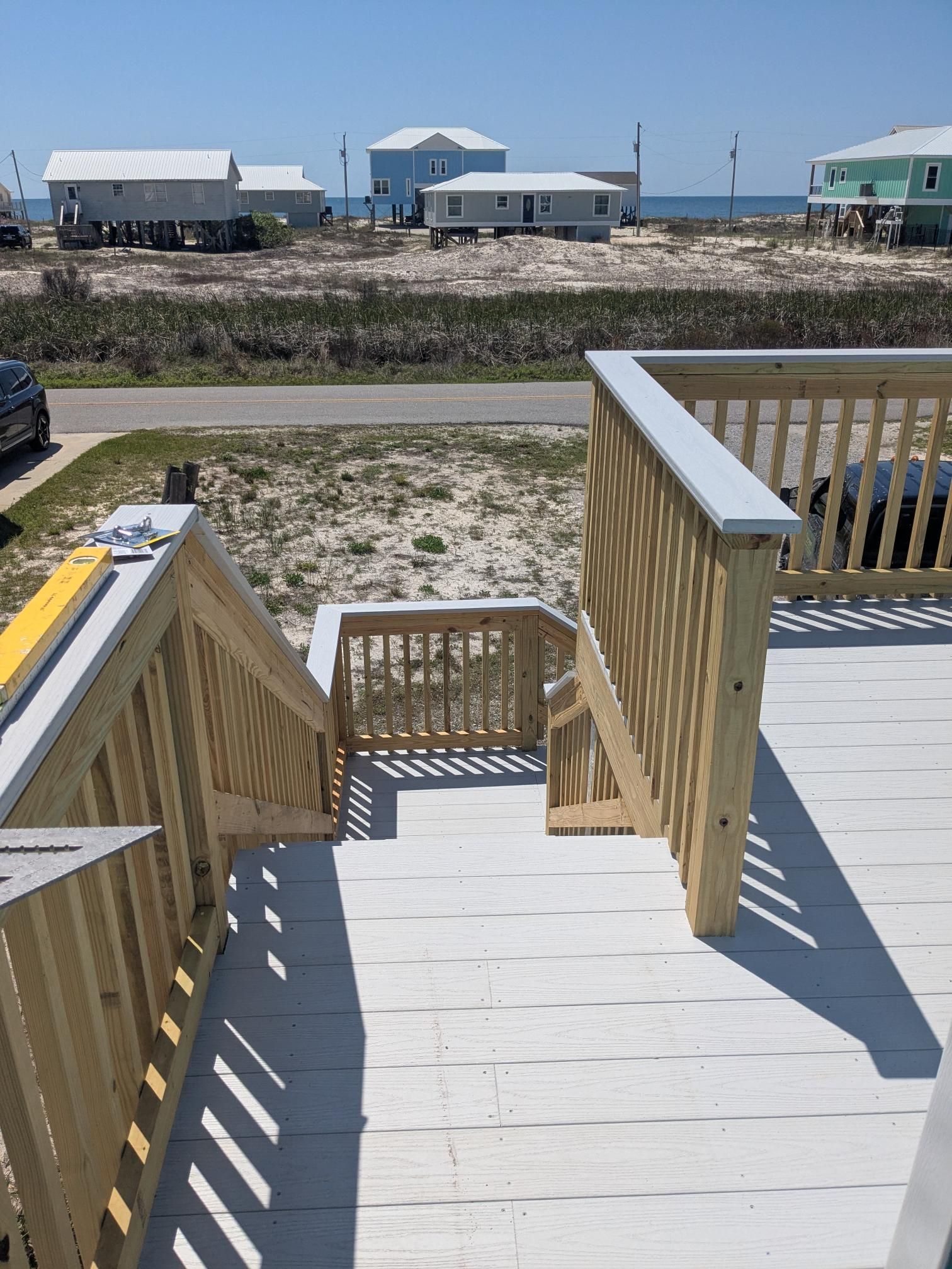Wooden deck with steps leading down to a sandy area; houses and the sea in the background.