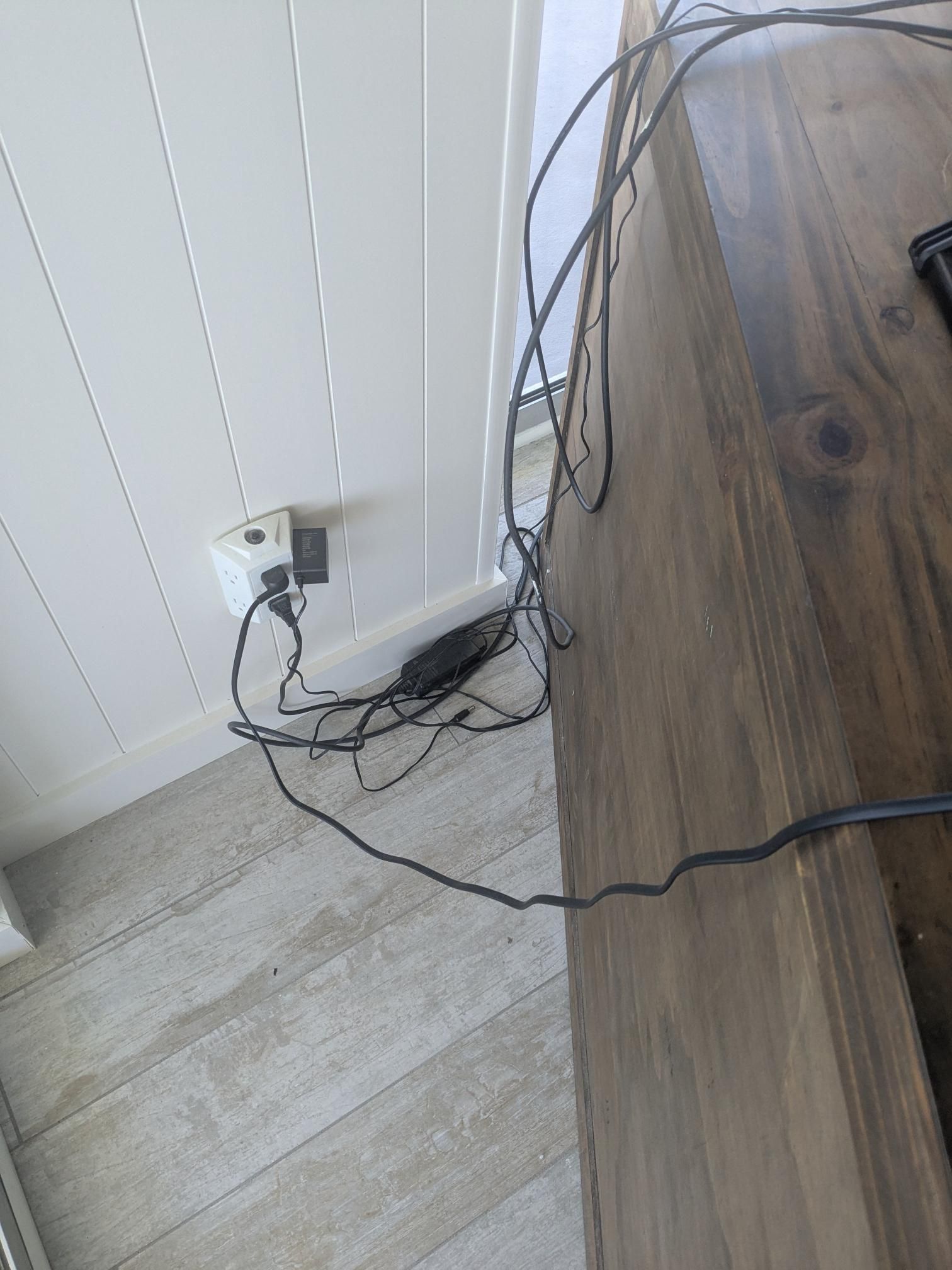 Electrical outlet with tangled black cords on a white wall near a wooden surface.