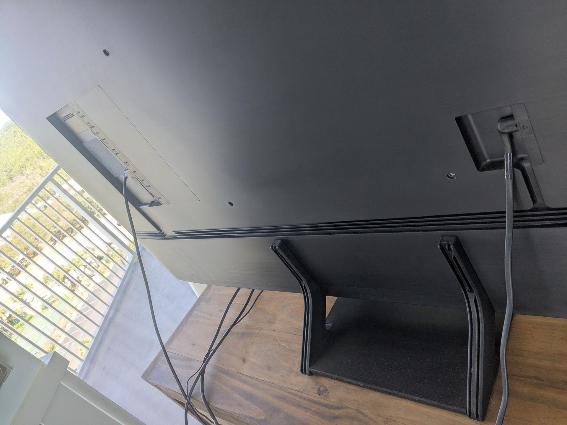 Backside of a dark TV with connected cables and a stand on a wooden surface, outdoor view in the background.