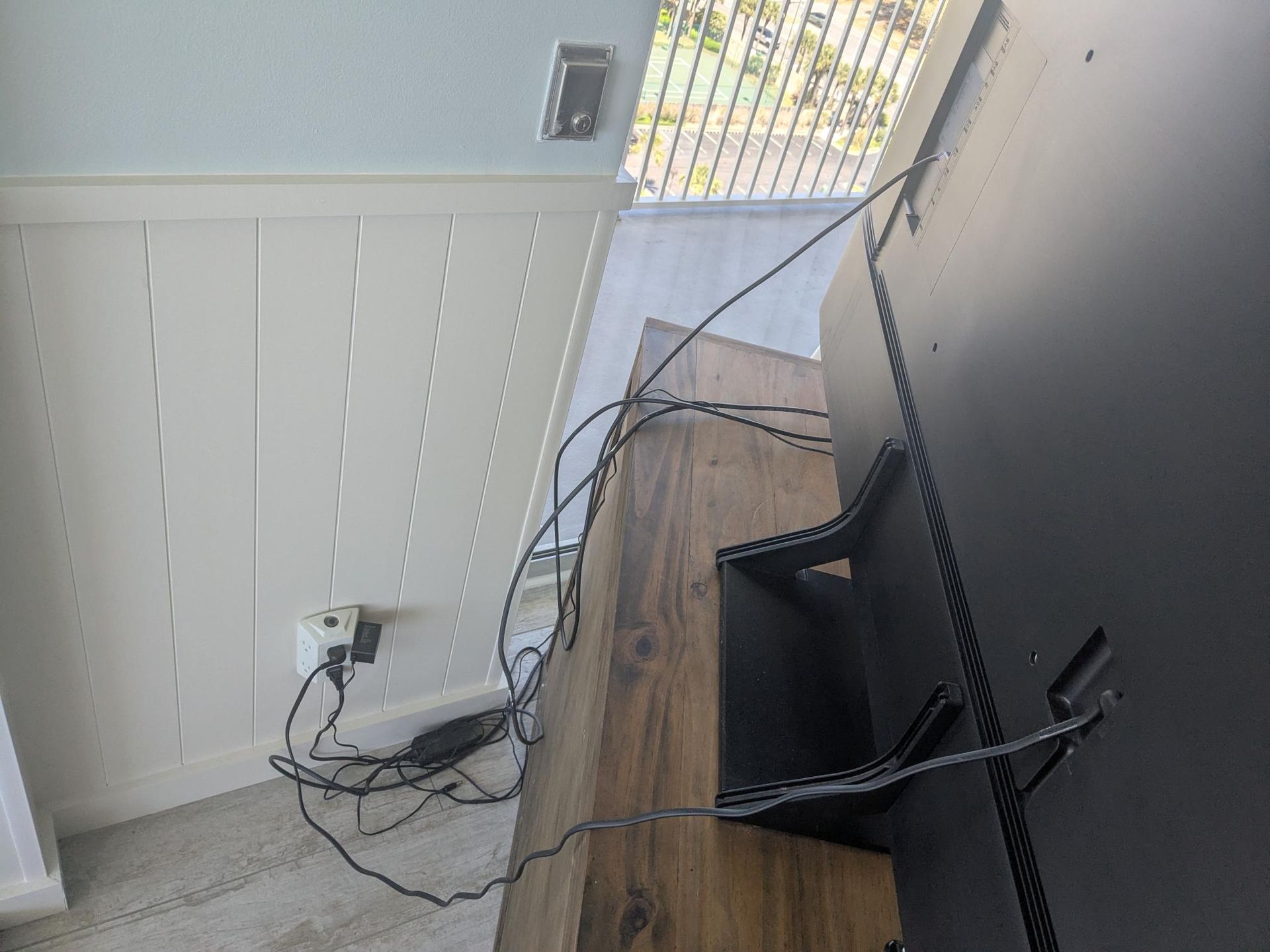 TV on wooden stand with tangled wires; white paneled wall, outlet, and window visible.