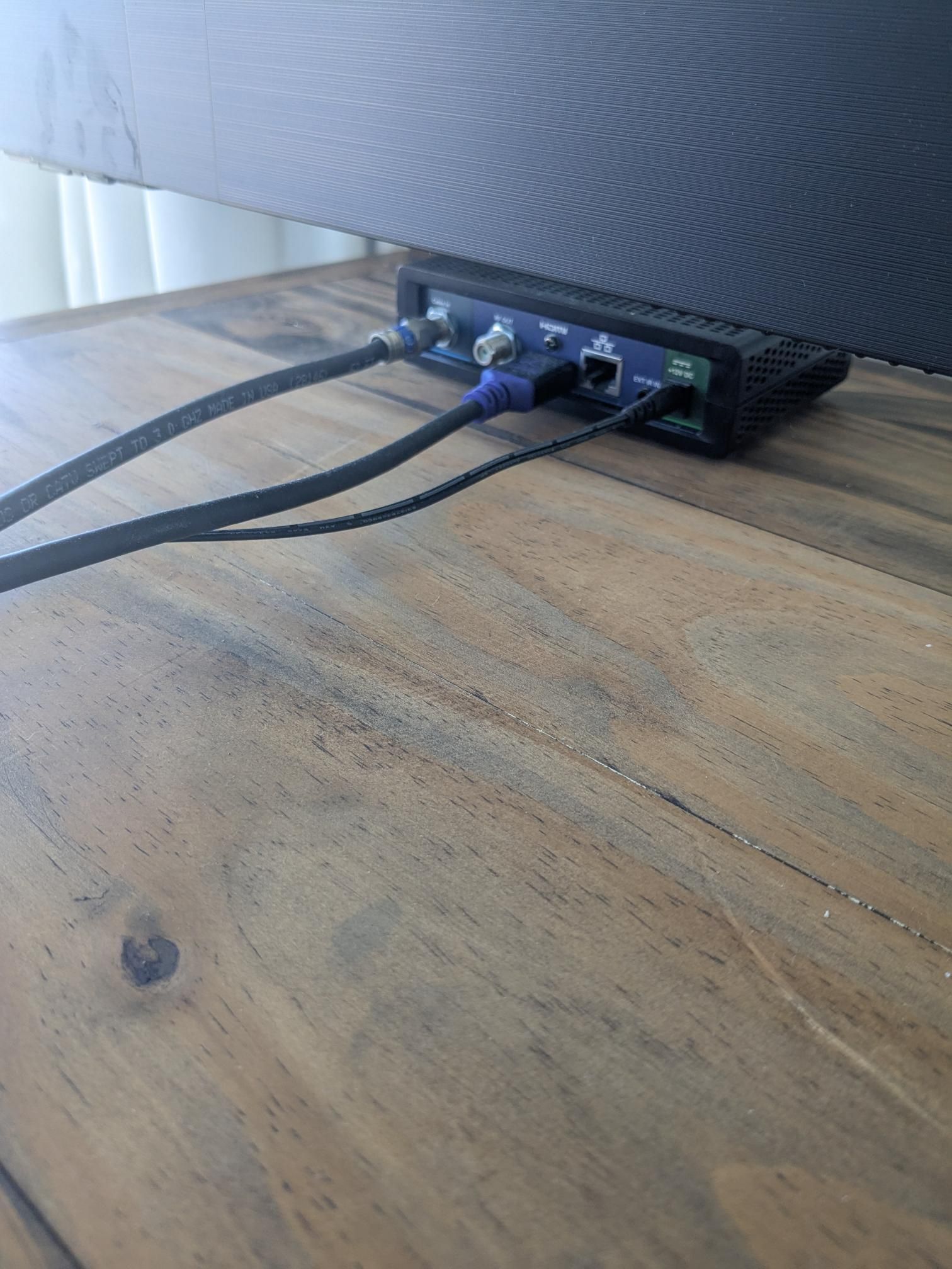 Black cables plugged into a black device under a television on a wooden table.