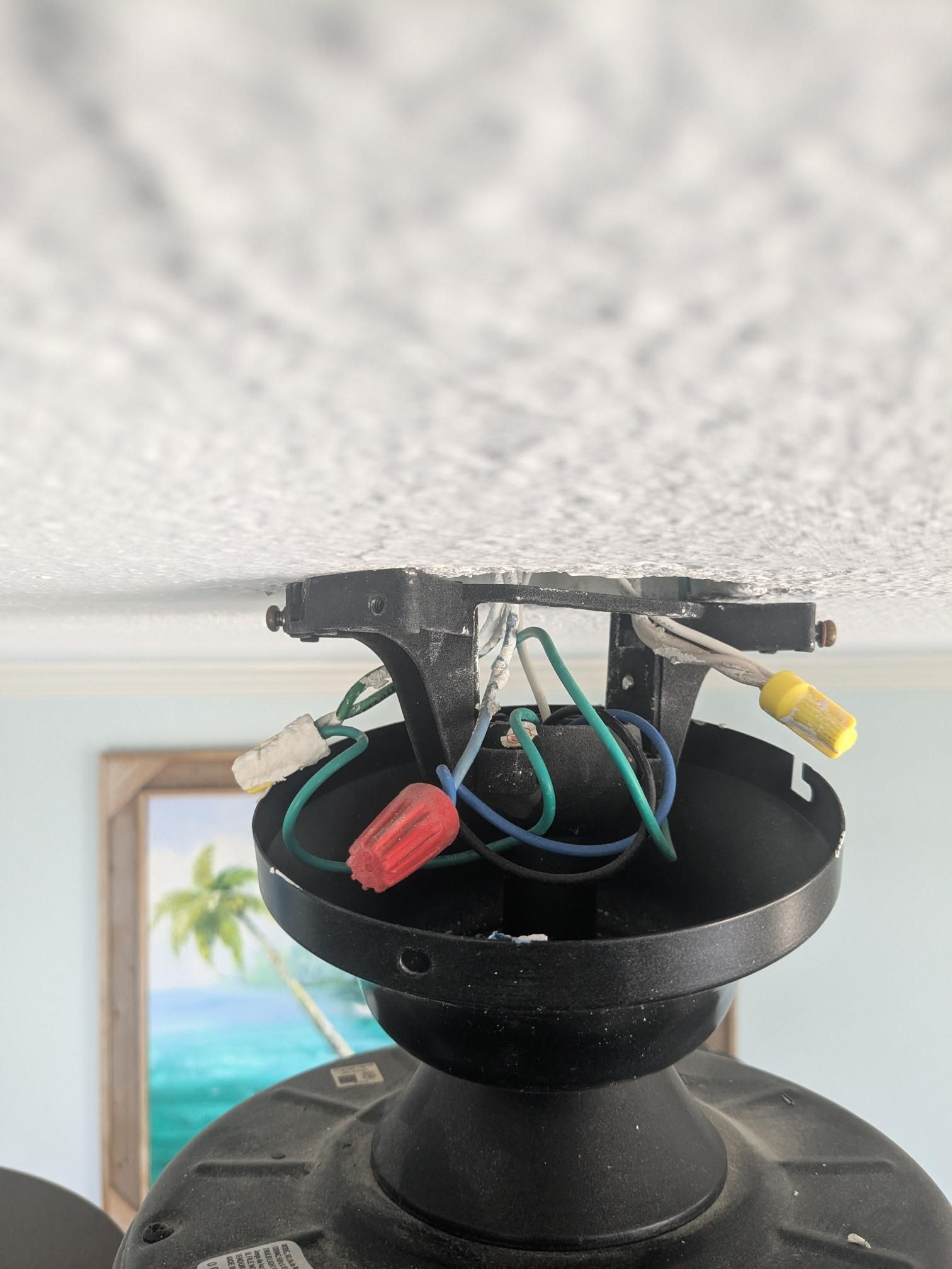 Close-up of a ceiling fan with exposed wires and a wire connector. The ceiling is white with texture.